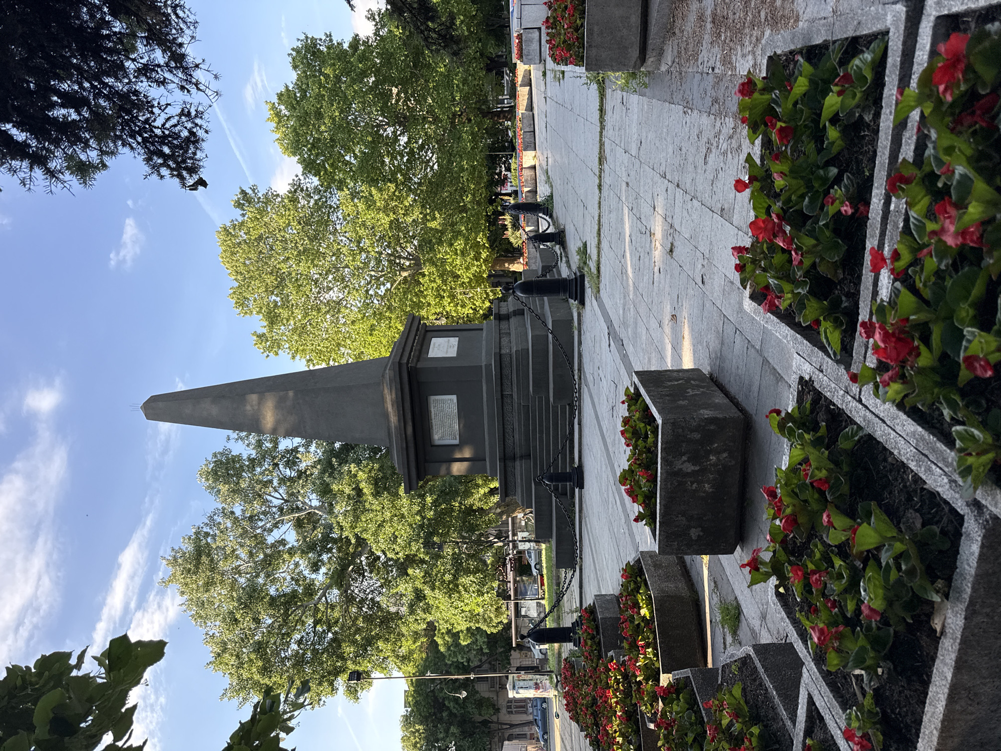 The image shows an obelisk monument in an urban setting, likely a park or memorial site. The obelisk is constructed of dark stone, featuring plaques and a stepped base surrounded by chains. It stands tall against a background of green trees and a blue sky with scattered clouds.
Adjacent to the monument is a paved walkway, lined with concrete planters filled with red flowers. The sidewalk extends into the distance, suggesting a well-maintained urban space. In the background, there is a street scene, with vehicles, street lamps, and other architectural structures visible.