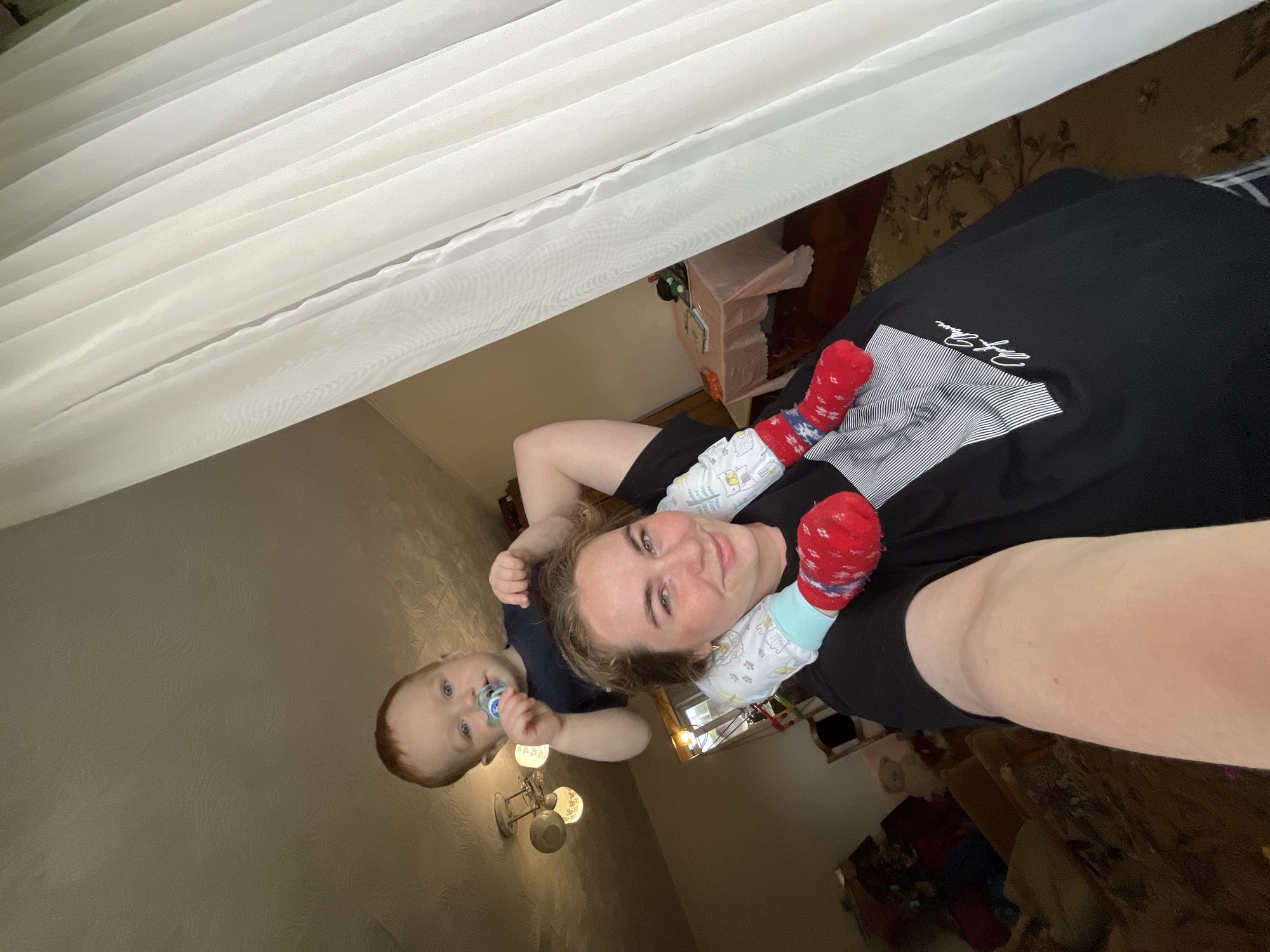An adult female, appearing in her 20s or 30s with light skin and light brown hair, captures a selfie while an infant, approximately 10-18 months old, sits on her shoulders. The female, smiling, wears a black t-shirt featuring a white geometric and script design on the left chest and a small gold hoop earring in her left ear. The infant, with light red-brown hair and blue eyes, looks directly at the camera, holding a blue and white pacifier in his mouth. He is dressed in a dark blue t-shirt, white full-body pajamas with light blue cuffs and a cartoon animal pattern, and red socks with a white speckled design.
The scene is set indoors, within a residential dwelling in Сосново, Russia. The background reveals light-colored, possibly beige, walls and a textured ceiling from which a multi-bulb chandelier hangs. A large window on the right is covered by multiple panels of sheer white curtains, providing natural light. To the immediate right in the background, a small table draped with a light pink tablecloth holds a stack of books and various small objects. To the left in the background, a section of a room with indistinct furniture and a teddy bear is visible. The floor is covered with a patterned rug.