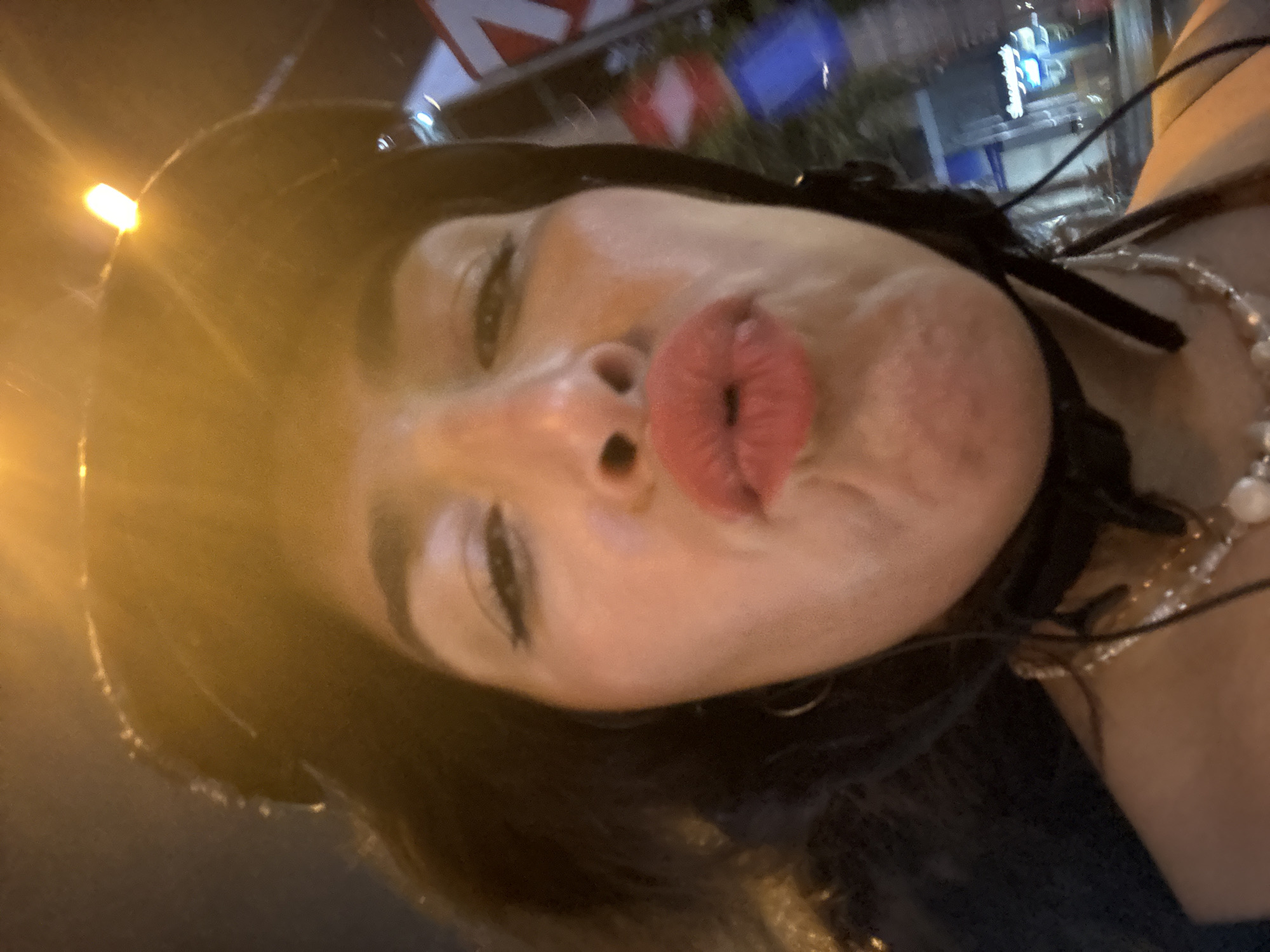In Thủ Đức City, Vietnam, a close-up, front-facing selfie is captured at night from the perspective of a person riding on a motorcycle. The subject, a woman with dark hair, is wearing a black helmet. Her face is illuminated by the glow of streetlights, which also create a bright light source at the top of the helmet. She is making a kissing gesture with bright red lipstick on her lips, with her mouth puckered and her chin slightly tucked. She is also wearing a layered necklace, with the top layer appearing to be a thin chain with small embellishments and the bottom layer featuring larger pearls. In the blurred background, elements of a city street are visible, including streetlights, buildings, and signage, suggesting an urban environment.