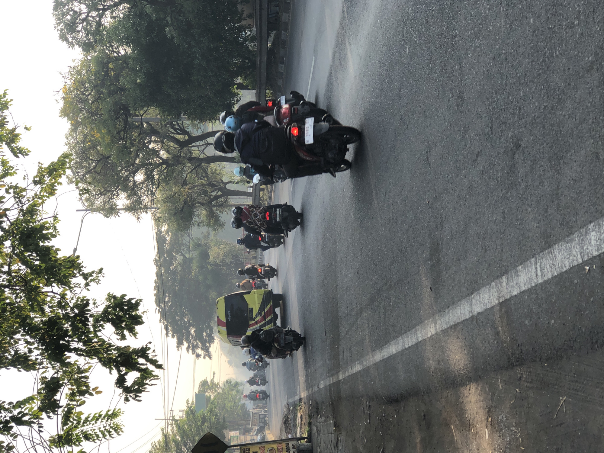 A group of motorcyclists ride in formation on a paved road in Kludan, Indonesia. The road is flanked by trees and foliage on both sides. In the background, a yellow van with red and black stripes is visible. The sky is hazy, suggesting it is either early morning or late afternoon. The motorcyclists are wearing helmets and riding in the same direction. The scene appears to be a typical commute in Indonesia, with many people relying on motorcycles for transportation. The presence of a yellow van and the tropical vegetation further indicate a location in Indonesia.