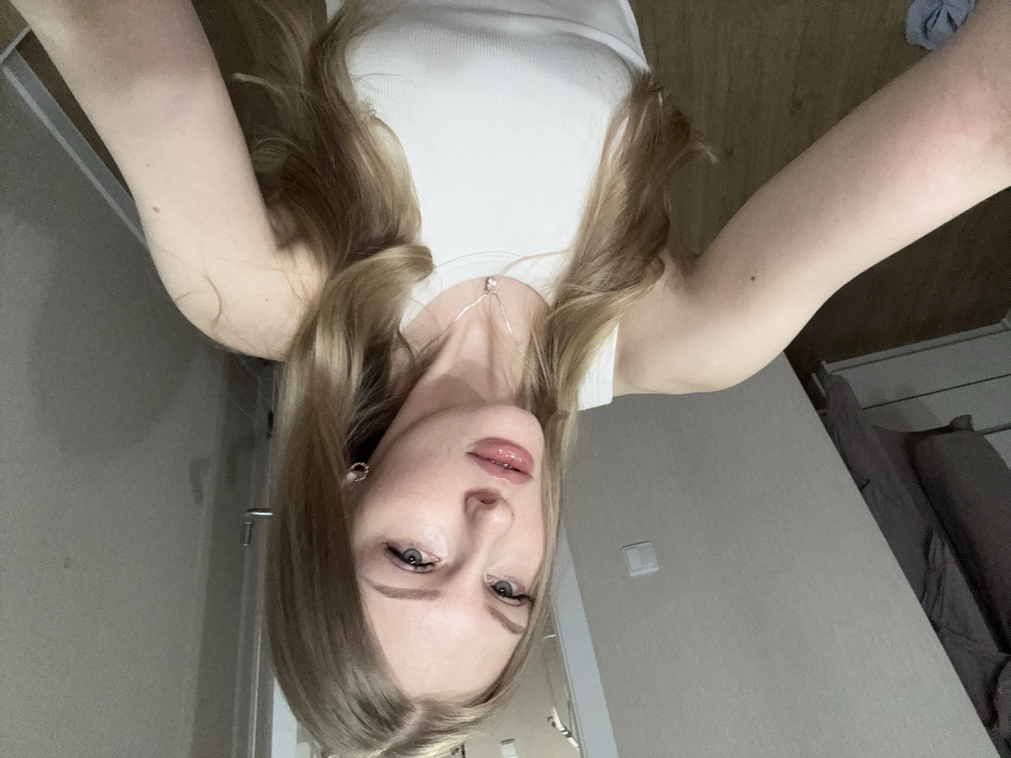 The image captures an upside-down selfie of a young woman taken indoors in Perm, Russia. The woman has long, wavy blonde hair, parted in the middle, and is wearing a simple white ribbed tank top. She has fair skin, light-colored eyes, and her lips are accentuated with a glossy pink shade. A delicate silver chain necklace with a small pendant rests on her chest. Small silver hoop earrings are visible on her ears. Her expression is neutral, and she makes direct eye contact with the camera, which she appears to be holding above her head.
The background consists of a light-colored wall, with a white light switch visible on the right. To the far right, a dark piece of furniture, possibly a bed or sofa with dark upholstery, is partially visible. The floor below appears to be a light wood or laminate. The overall lighting is soft and even, characteristic of an indoor setting. No discernible text is present in the image.