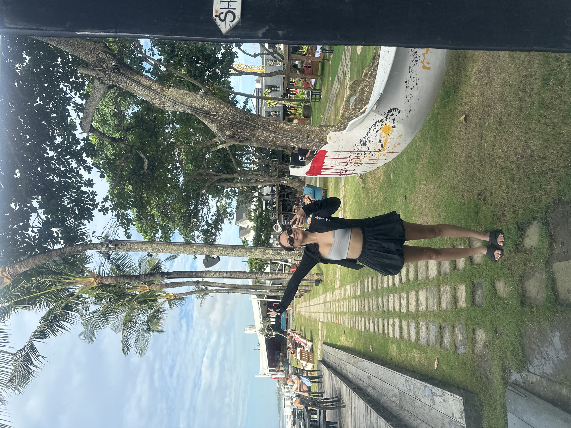 The image shows a person taking a selfie in North Kuta, Indonesia. The individual, wearing a crop top, black jacket, and skirt, stands on a stone and grass path. In the background, there is a tree, and a white structure with colorful splatters can be seen. Further in the background, there are people sitting at tables, suggesting a restaurant or outdoor seating area. Palm trees and a partly cloudy sky are visible. The image is taken from under a structure, creating a frame around the scene.