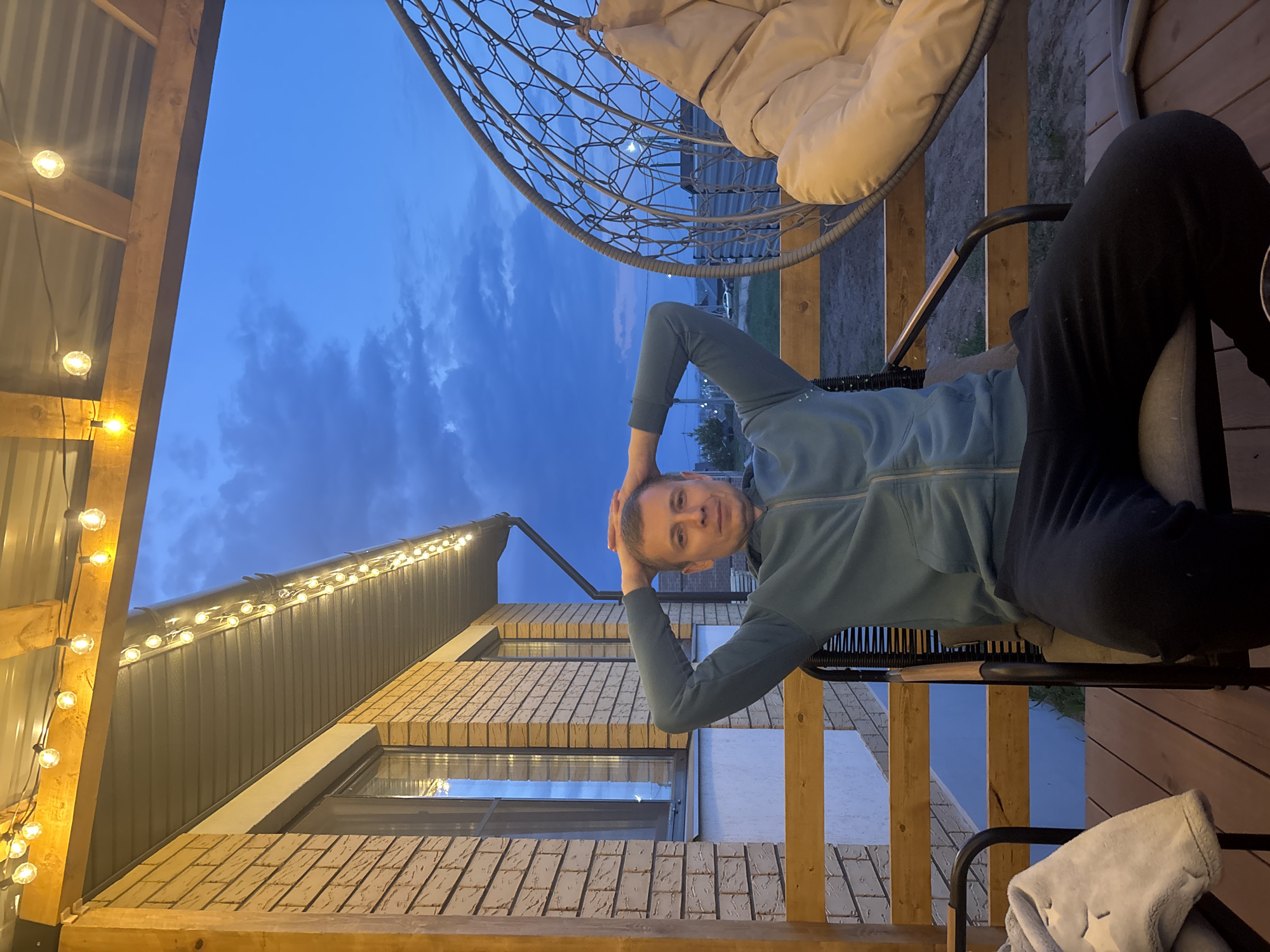 A person is sitting in a chair on a wooden deck. They are wearing a blue sweatshirt and dark pants, with their hands behind their head. Above and behind them is a hanging chair with a large cushion. In the background is a brick building with lit windows and a roof adorned with string lights. The sky is a dusk blue. Based on prior knowledge, this image was captured in or near Chaikovsky, Russia.