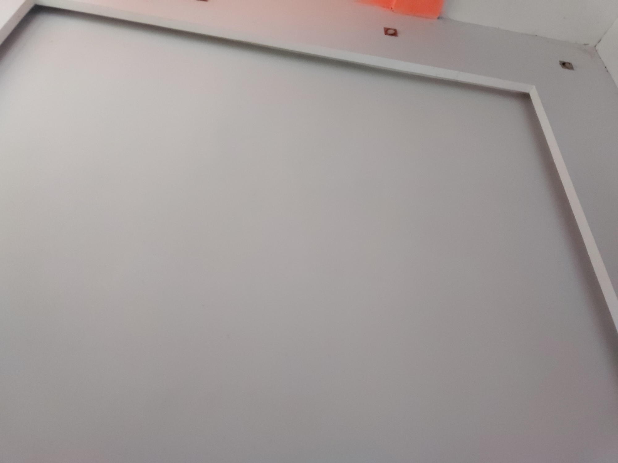 The image displays a static, indoor scene featuring a predominantly light gray wall. A white, rectangular trim frames a large central section of this wall, creating a clean, defined boundary. Above this trim, closer to the ceiling, several details are visible: an orange-colored object is partially seen in the upper left corner, and two small, dark square-shaped fixtures or cutouts are present against the white ceiling or upper wall area. One of these dark squares is located above the orange object, while the other is near the upper right corner of the frame. The overall aesthetic is minimalist, dominated by neutral tones of light gray and white, with small accents of orange and dark brown/black from the fixtures. No individuals are present, and no activity is taking place. The lighting appears even, suggesting a well-lit interior space. There is no visible text within the frame. The location is Santa Ana, Venezuela.