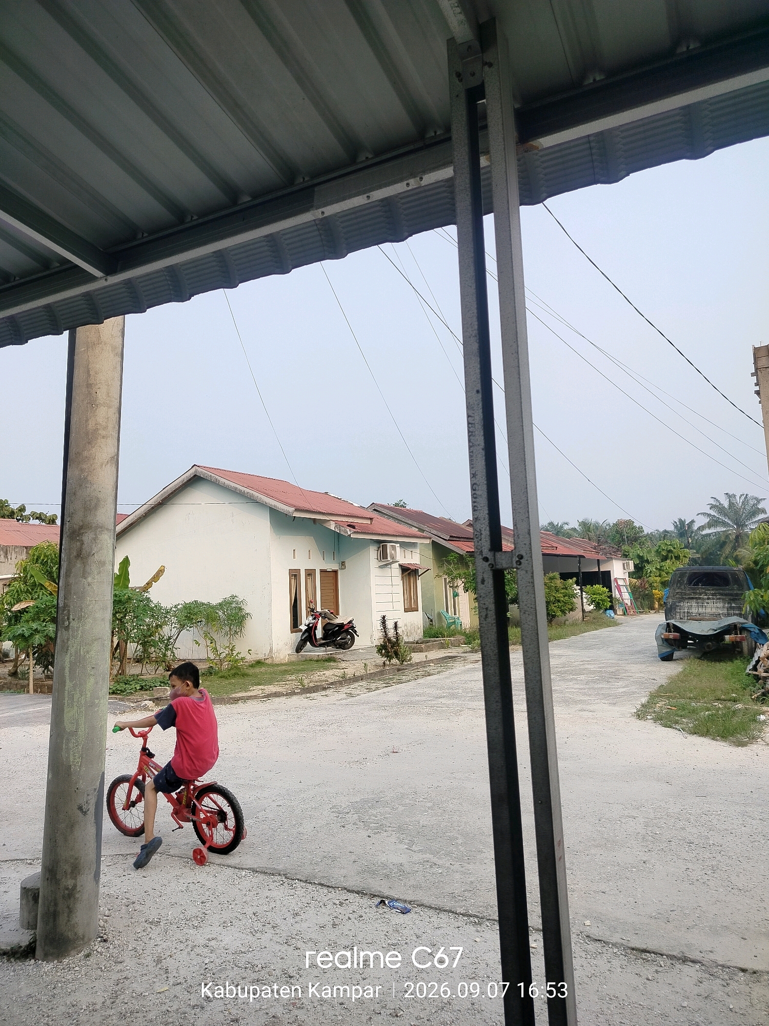 A young boy in a red shirt and dark shorts rides a red bicycle with training wheels on a gravel road in a residential area. The boy is positioned on the left side of the image, facing right. The bicycle has black tires and red rims. In the background, there are several houses with red roofs, some with white walls and brown window frames. A motorcycle is parked in front of one of the houses. A blue pickup truck is visible further down the road on the right side. Lush green foliage and palm trees are scattered throughout the scene. Overhead, a corrugated metal roof structure is visible in the foreground, supported by metal beams. The sky is overcast and pale blue. The timestamp on the image indicates it was taken in Kabupaten Kampar, Indonesia, on September 7, 2026, at 16:53.