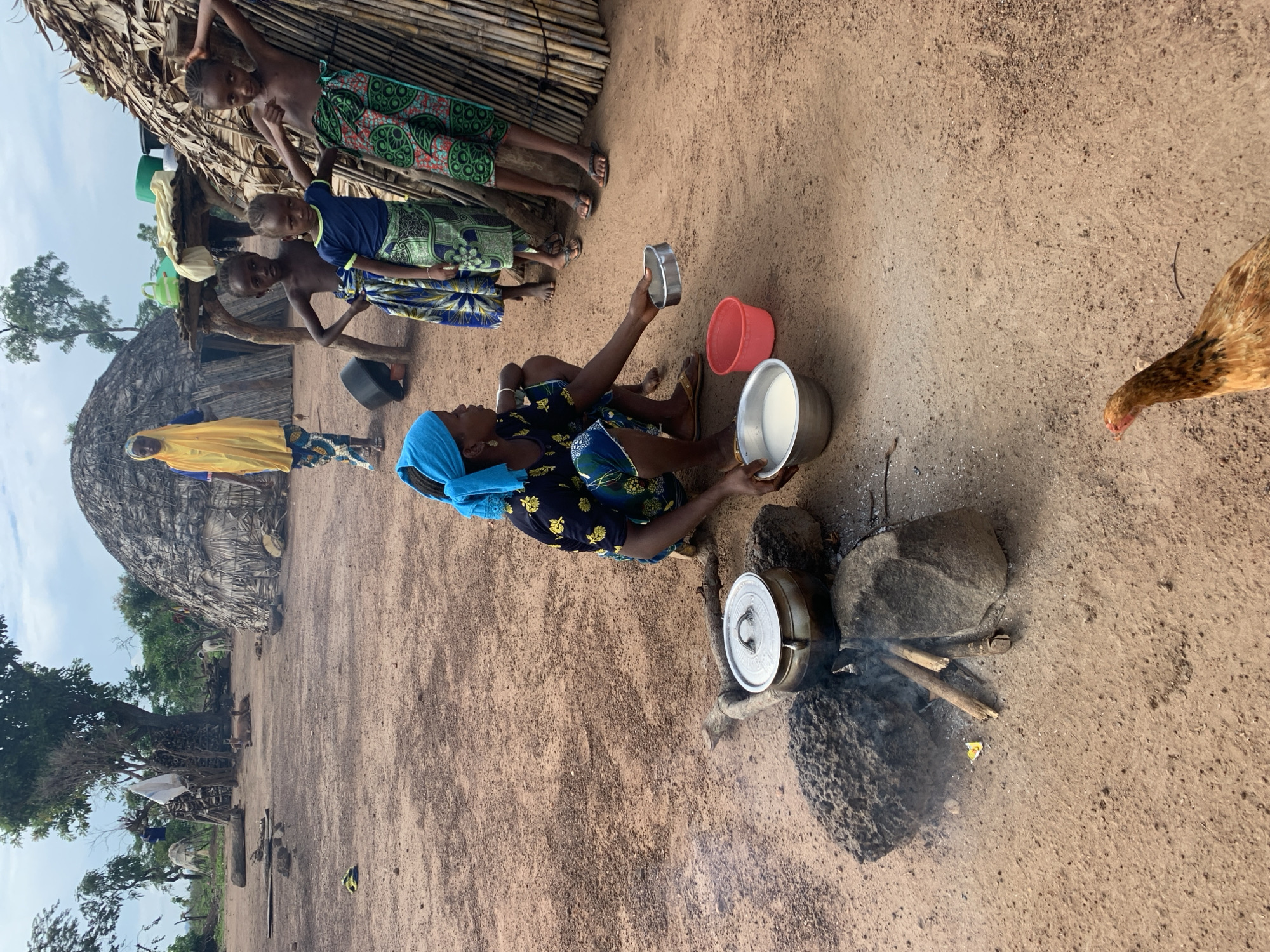 An outdoor scene in Nandu, Nigeria, depicts a village setting. In the foreground, a woman wearing a blue headscarf and a patterned dress crouches next to a cooking fire. A metal pot sits on rocks over the fire, with smoke rising. She is holding a small metal bowl and a larger metal bowl containing a white liquid, with a red bowl nearby. Several children are visible in the midground, some standing near a thatched structure and others in the background. One child wears a yellow garment. The ground is sandy and dry, with several thatched huts and trees scattered throughout the landscape. A chicken is present in the lower right corner of the image.