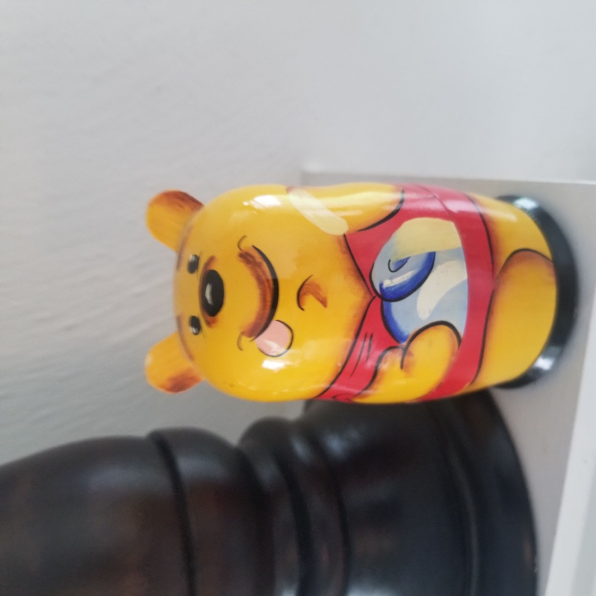A Winnie the Pooh nesting doll is displayed on a white shelf in a room. The doll is primarily yellow with a red shirt and blue and white accents on its chest. The doll's face is painted with a smiling expression, tongue sticking out, and rosy cheeks. To the left of the doll is a dark, curved object, possibly a lamp base or part of a piece of furniture. The background consists of a white wall and the white shelf on which the objects are placed. The image appears to be taken in a domestic setting. The location of Virginia Beach, United States of America, is not discernible from the visual information in the image.