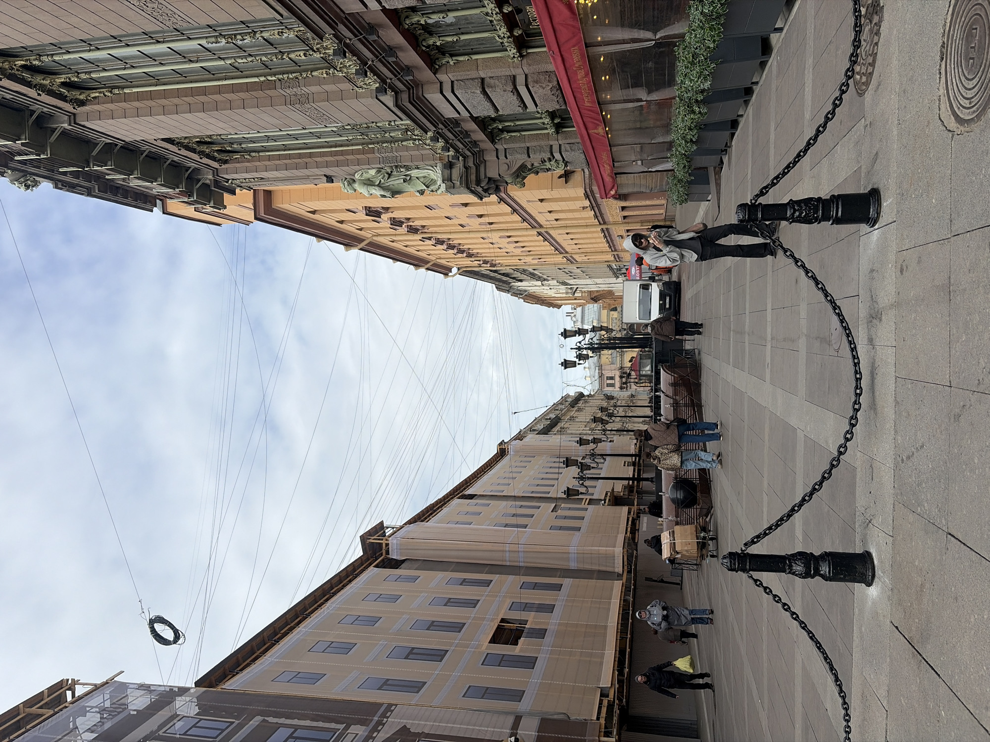 The image depicts a street scene in Saint Petersburg, Russia. The street is lined with buildings on both sides. The building on the left is covered in scaffolding and protective screens, indicating ongoing construction or renovation. The building on the right exhibits ornate architectural details, including statues and decorative elements. A red awning is also visible on this building. Overhead, numerous electrical wires crisscross the sky.
Several people are present on the street, including individuals walking and standing. One person near the foreground is leaning on a black bollard with chains attached, possibly posing for a photo.
The street surface appears to be paved with stone. A manhole cover is visible on the right side of the street. Street lamps are also present along the street.