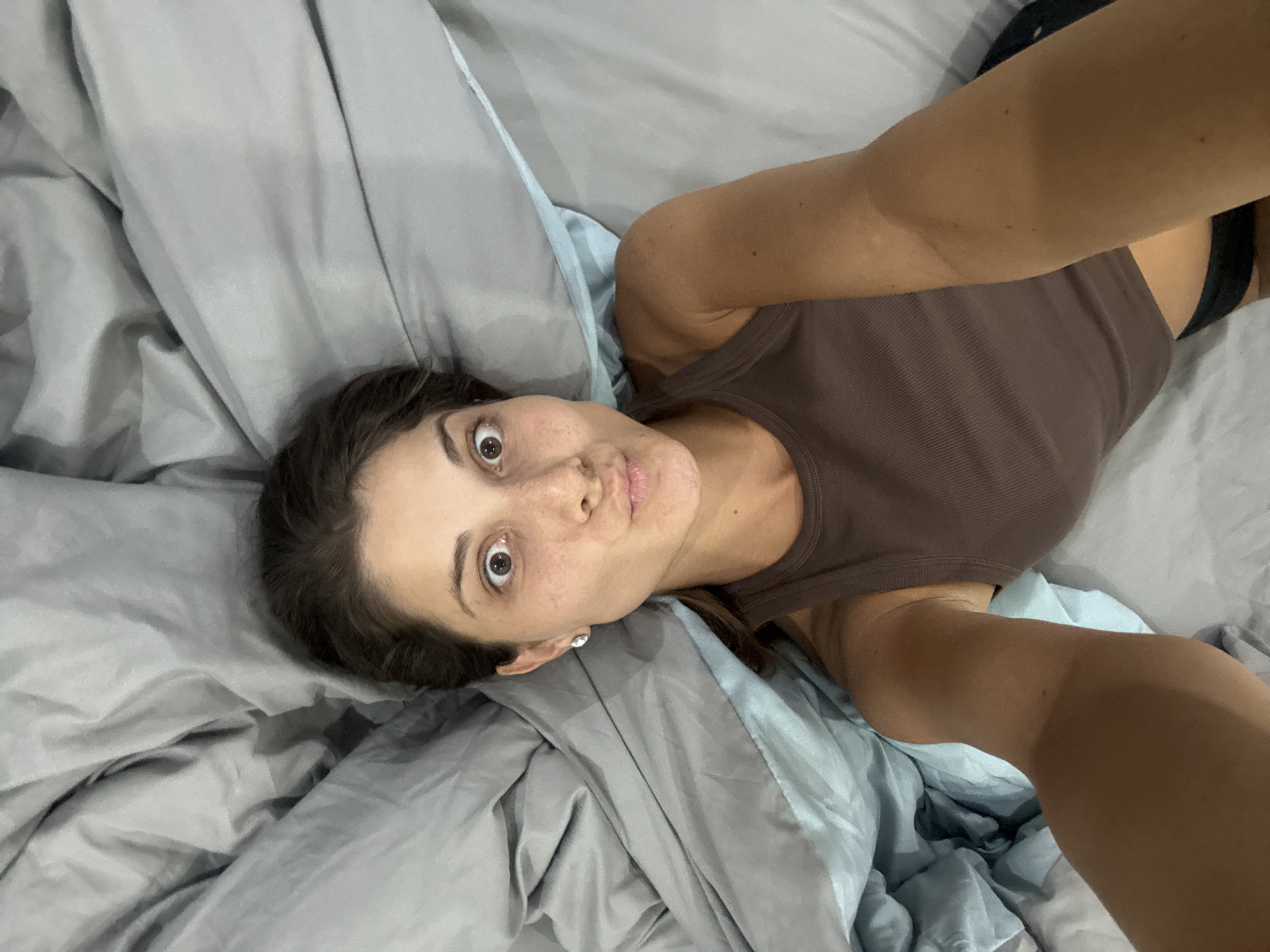 A young woman is captured in a selfie, lying on what appears to be a bed covered with rumpled grey and light blue bedding. She has brown hair, visible freckles on her face, and dark eyes that are wide open, conveying a look of surprise or playfulness. Her lips are slightly puckered in a gentle pout. She is wearing a simple brown ribbed tank top, and a black strap, possibly from a bra or another undergarment, is visible near her right shoulder. A small, shiny stud earring is visible in her left ear. Her arms are extended, holding the camera to take the overhead shot, and her tanned skin is visible. The lighting is soft and even, suggesting an indoor setting without strong natural light. There are no discernible location cues within the image itself, though the context provided indicates the location is Đà Nẵng, Vietnam. No text is visible in the image.