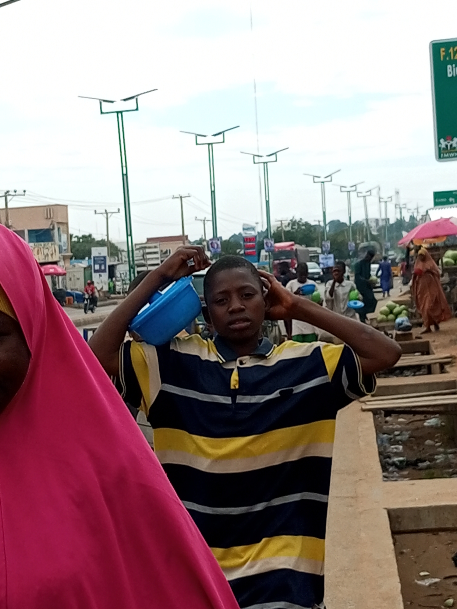 This image depicts a street scene in Gombe, Nigeria. In the foreground, a young boy wearing a horizontally striped t-shirt (dark blue, yellow, and white) is holding a blue plastic container on his shoulder and has his right hand touching his head. To his left, a person wearing a bright pink hijab is partially visible.
The background shows a busy street with several lamp posts, some of which appear to be solar-powered with their distinctive panel design. There are also other people visible, some carrying containers, and market stalls with produce like watermelons. Vehicles, including cars and a motorcycle, are present on the road. A green sign with white lettering, partially obscured, is visible on the right side of the image. The overall environment suggests a bustling urban marketplace or street.