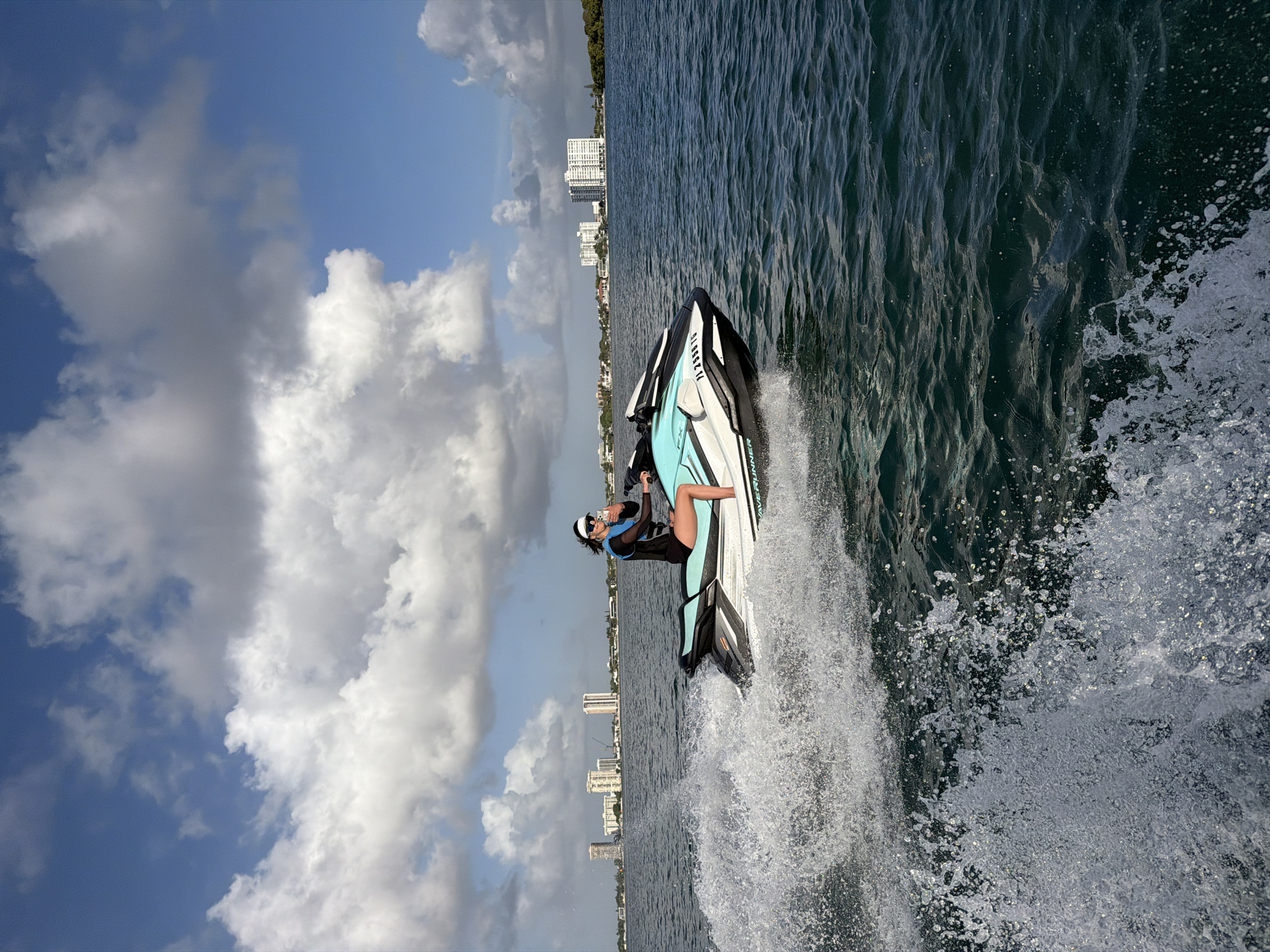 A medium outdoor shot portrays a person riding a jet ski on the water. The jet ski is predominantly white and black with light blue accents. The rider is dressed in dark attire and sunglasses. They are positioned on the jet ski with the skyline of North Miami, Florida, including high-rise buildings, visible in the background. The sky features scattered clouds. Water spray is prominently visible around the jet ski.