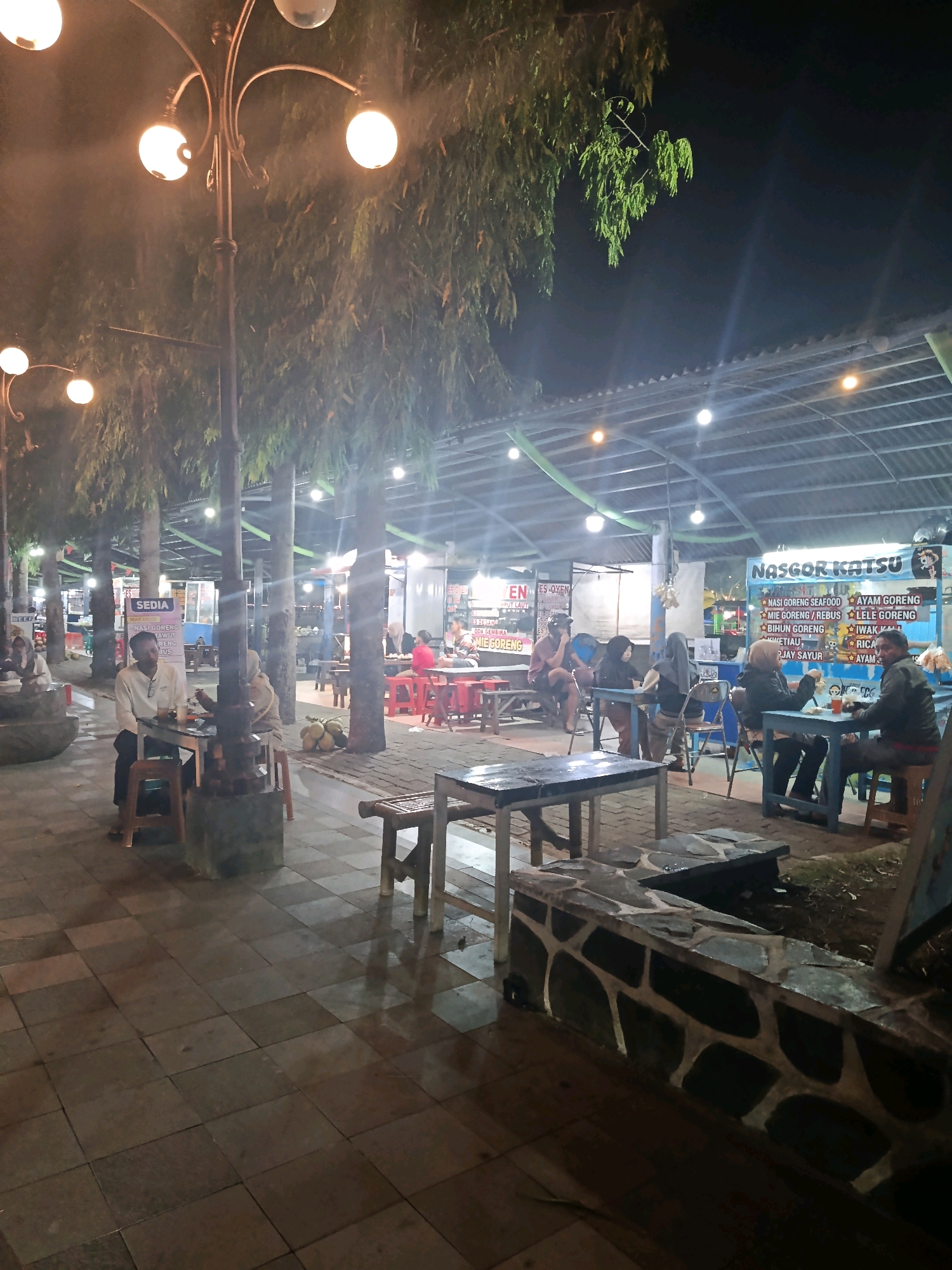 This image depicts an outdoor dining area at night in Pacitan, Indonesia, likely a food court or a row of food stalls. A prominent street lamp with multiple globes illuminates the foreground. Several people are seated at tables, some eating and others conversing. The tables are a mix of metal and wooden designs, with some set on a tiled patio. In the background, food stalls are visible, with signage indicating food items like "NASGOR KATSU" (Fried Rice Katsu), "AYAM GORENG" (Fried Chicken), and "LELE GORENG" (Fried Catfish). The area is covered by a corrugated metal roof supported by posts, and the overall ambiance is that of a casual, communal dining space. The lighting suggests artificial illumination is the primary light source.