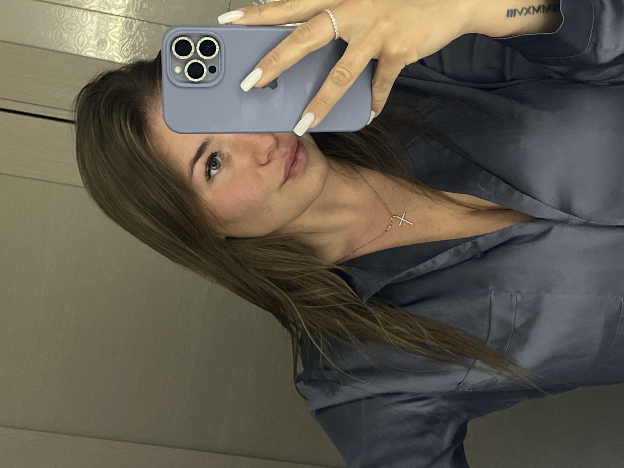 A woman takes a selfie in a mirror. She holds a lavender-colored smartphone with a triple-lens camera. The phone covers part of her face, but her left eye, nose, and lips are visible. She wears a silver cross necklace and a ring on her ring finger. Her hair is brown and straight. She wears a navy blue button-down shirt or pajamas. A tattoo is visible on her inner arm. The background appears to be a wall with a textured surface. The photo does not contain enough information to accurately specify the location in the city of Budva, Montenegro.
