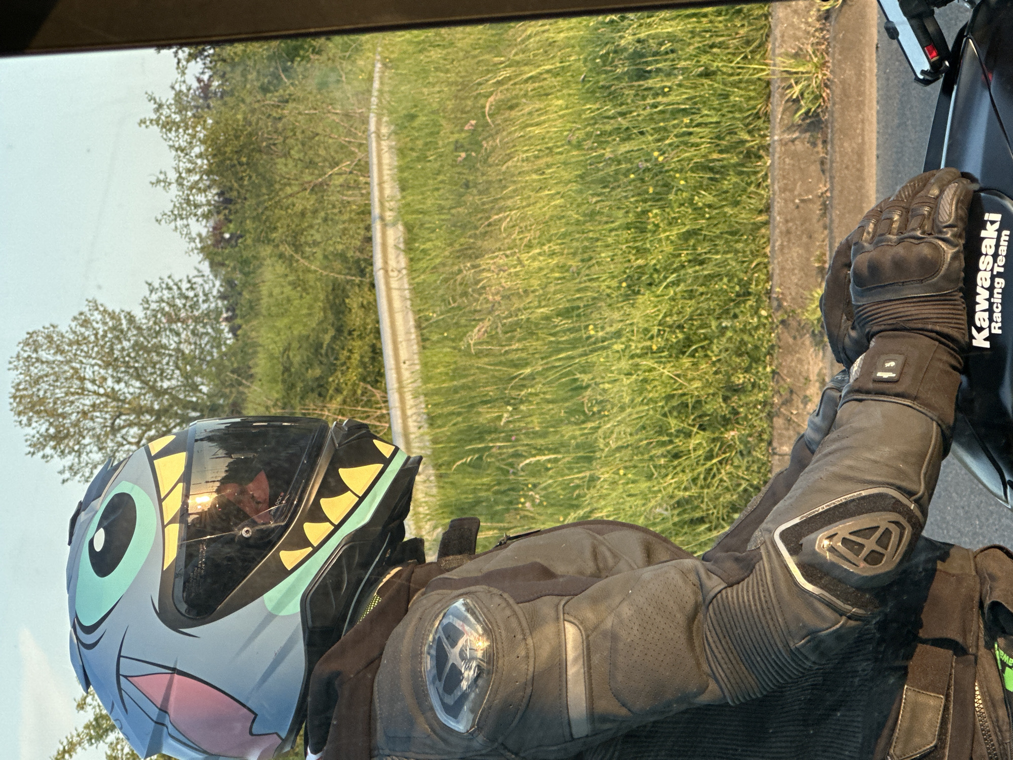 A person wearing a motorcycle helmet and gear is on a motorcycle in Villebon-sur-Yvette, France. The helmet has a cartoon design. The person is wearing black motorcycle gloves and a black leather jacket with protective padding. The motorcycle is black with "Kawasaki Racing Team" branding. The background shows a grassy area with trees and a roadside barrier.