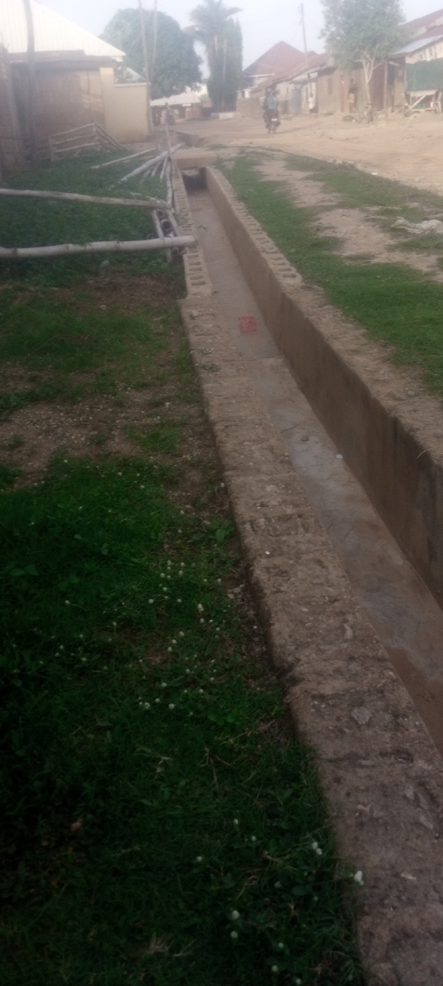 The image depicts a scene in a residential area. A concrete drainage ditch runs along the side of a road. On one side of the ditch is a grassy area with some white wildflowers. On the other side is a dirt road with houses visible in the background. A person on a motorcycle is riding down the road. Bamboo poles lay on the grass near the ditch. The location is labeled as Unknown, Unknown.