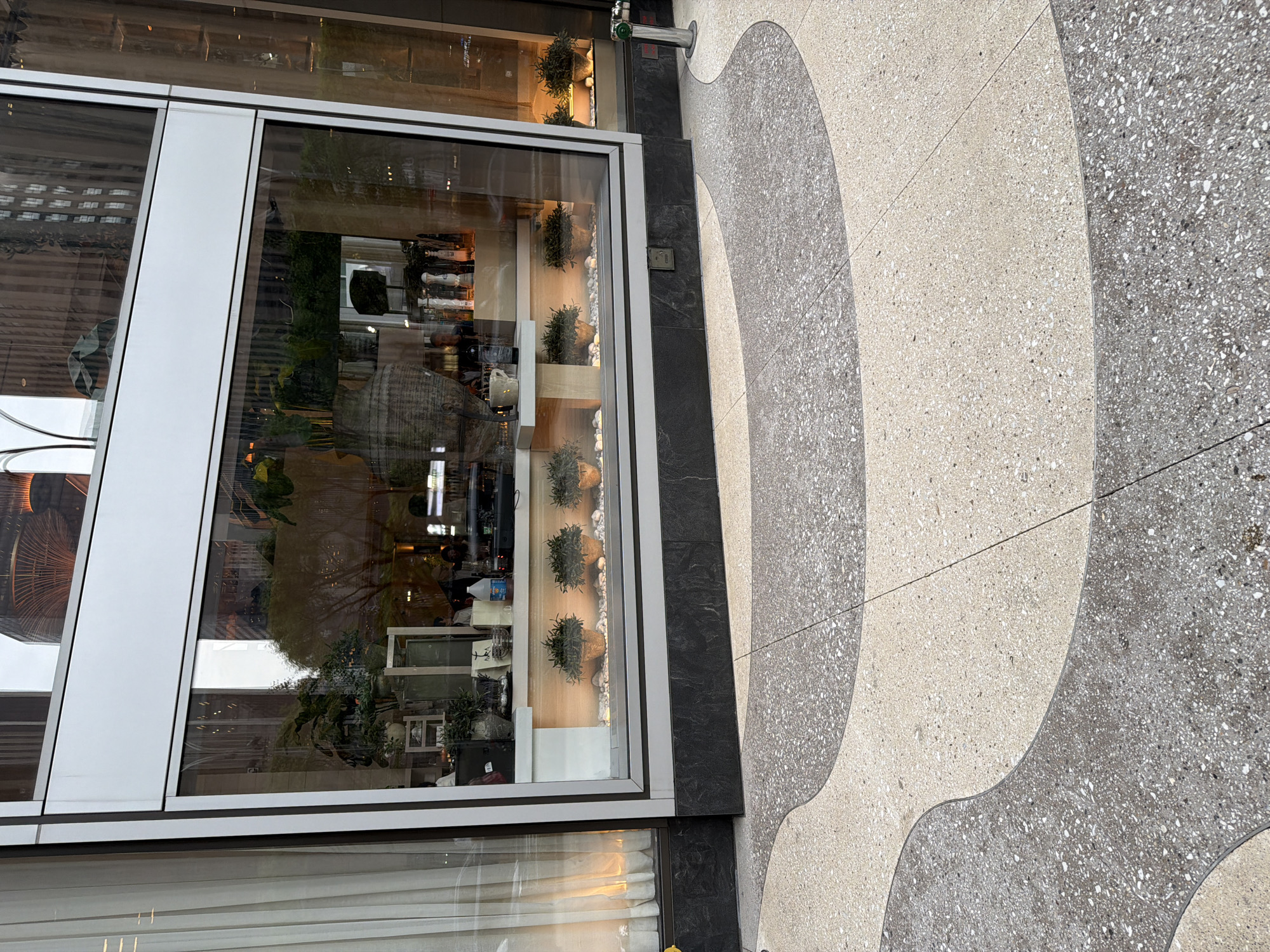 The image depicts a sidewalk in New York City, adjacent to a building with a large glass window. The sidewalk has a decorative pattern of light and dark gray terrazzo in curved shapes. A fire hydrant is visible near the building. The window reflects the surrounding buildings and streetscape, and displays decorative plants and interior objects on shelves. People are visible inside the building, blurred due to the reflection.