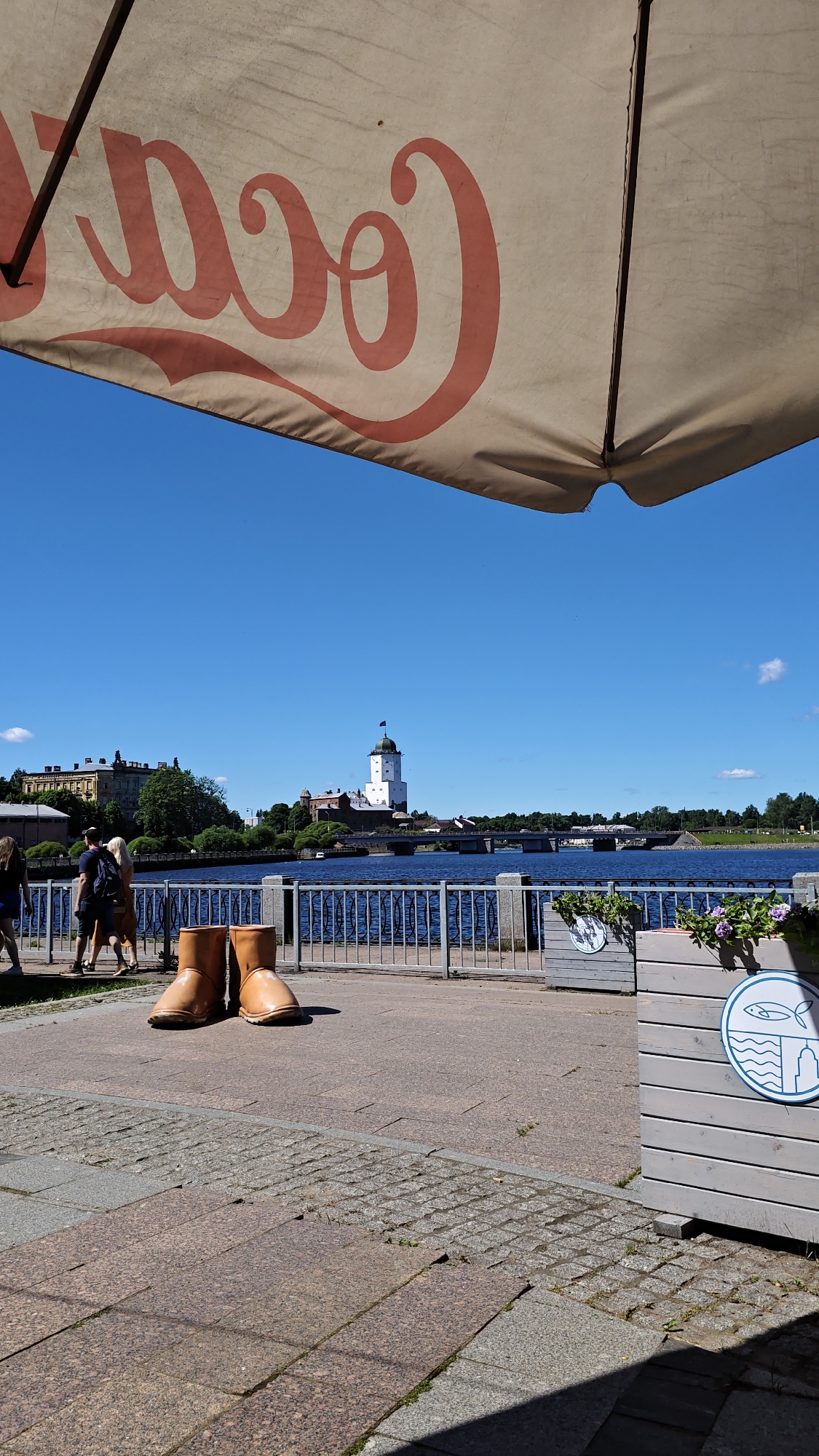 An outdoor scene captured on a sunny day features a view of Vyborg Castle in Vyborg, Russia. In the foreground, under a large umbrella with a faded Coca-Cola logo, a pair of oversized, tan boots are positioned on a cobblestone walkway. To the left, several people are walking along the promenade. A metal railing separates the walkway from a body of blue water. Beyond the water, Vyborg Castle, a white medieval structure with a dark-capped tower, stands prominently against a clear blue sky. A bridge spans the water in front of the castle, and buildings and trees line the distant shore. To the right, a wooden planter box decorated with a blue circular emblem featuring a fish and waves is visible. The pavement consists of large granite slabs and smaller cobblestones.