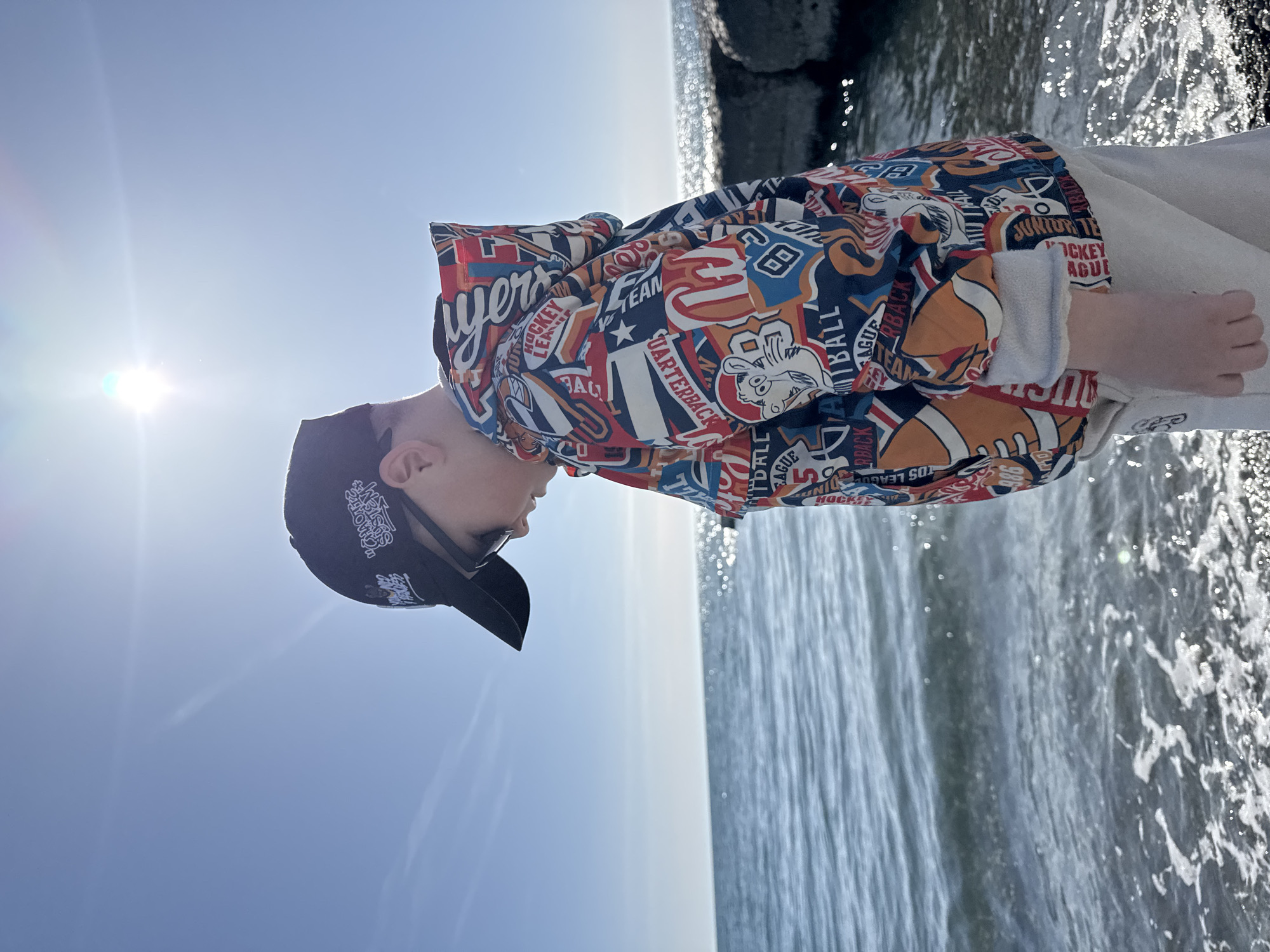 A person is depicted outdoors in a side profile, standing by a body of water, likely the Black Sea in Sochi, Russia. The bright midday sun is visible in the upper left, casting a strong flare and illuminating the scene under a clear blue sky.
The individual wears a dark baseball cap with white text, partially deciphered as "SMOKES SOCIETY," and dark sunglasses. They are dressed in a brightly colored hoodie or jacket covered in various sports-themed graphics and text. Visible phrases on the jacket include "PLAYERS," "DHOCKEY LEAGUE," "TEAM," "QUARTERBACK," "FOOTBALL," and "JUNIOR LEAGUE," featuring motifs of sports balls and team logos in red, blue, orange, and white.
To the right of the person, the sun-dappled surface of the water glistens, with a dark, rocky or concrete structure (possibly a breakwater) visible along the waterline in the background. The scene suggests a relaxed moment on a sunny day by the coast.