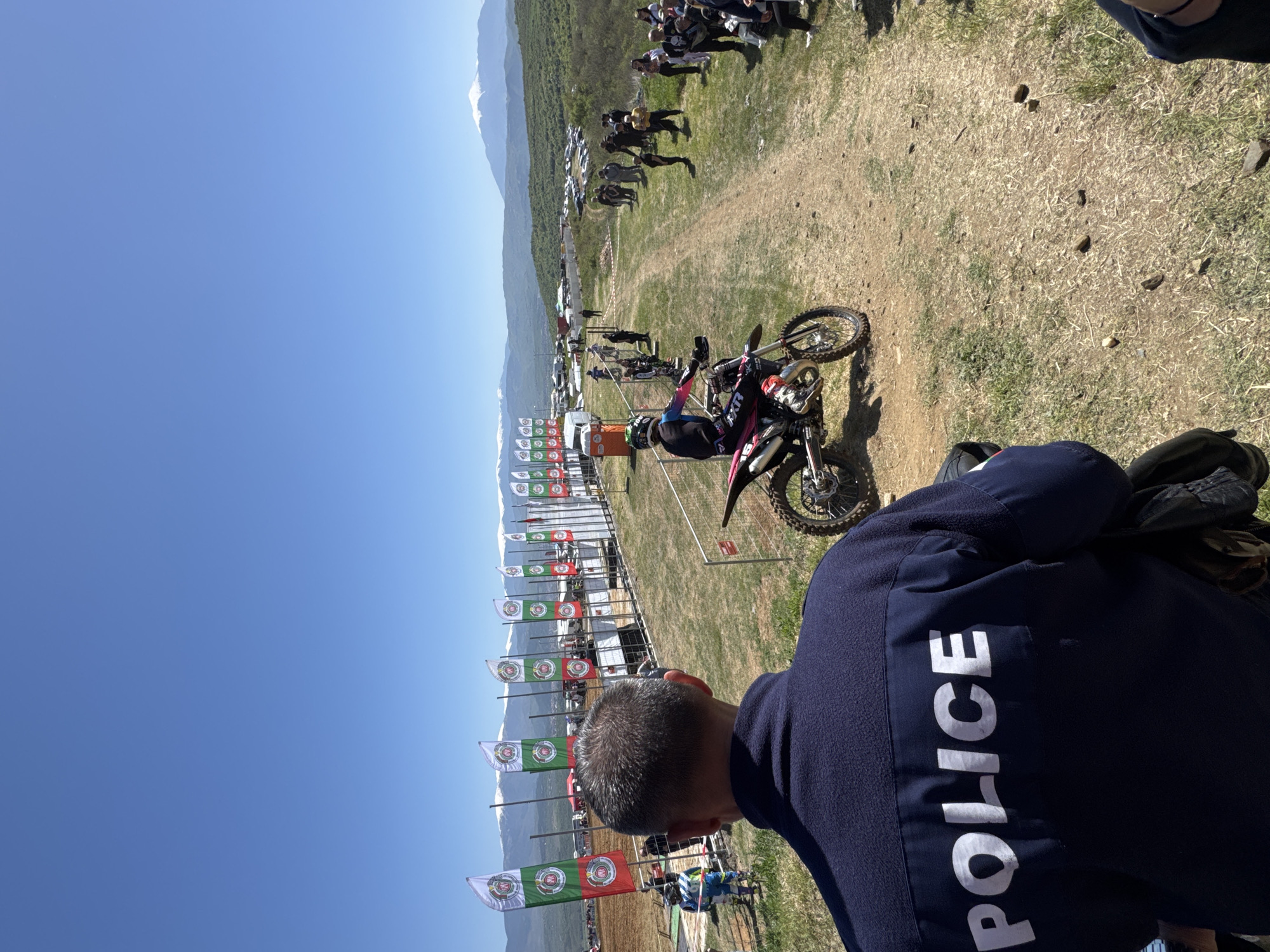 The image shows an outdoor motocross event in Selishte, Bulgaria, under a clear blue sky. A motorcycle rider wearing protective gear is paused on the track. A person in a police jacket stands in the foreground, facing the scene. Rows of flags representing the event or sponsor are visible along the side of the track. Spectators are visible in the background. Distant mountains add to the backdrop.