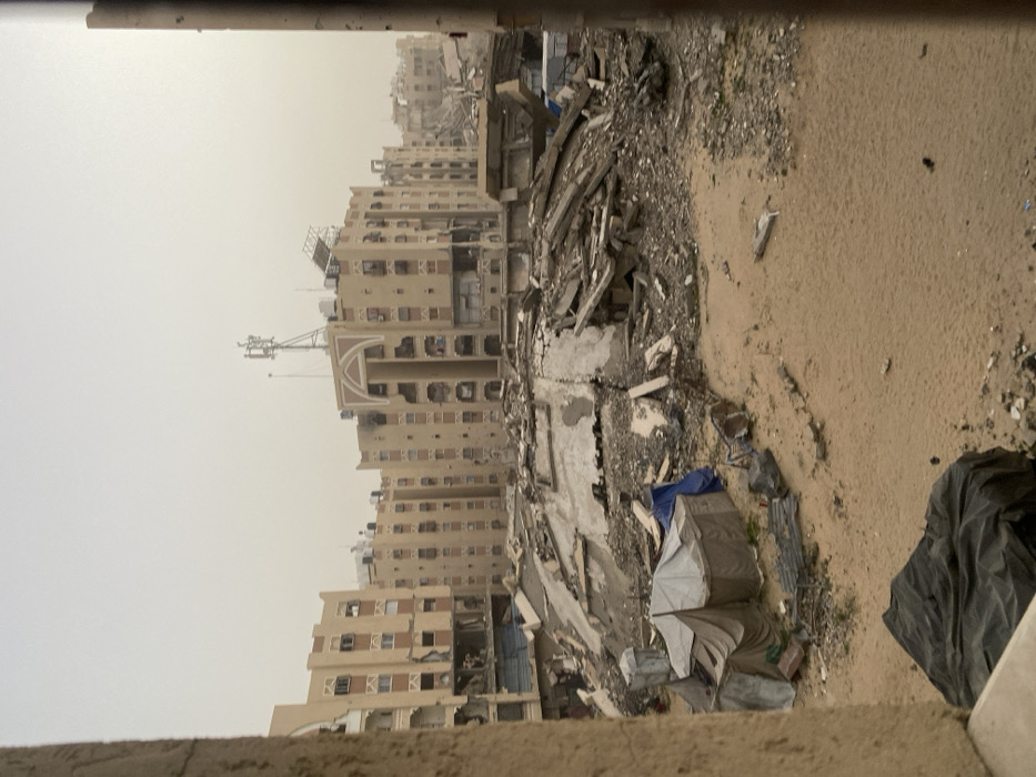 Destroyed buildings dominate the skyline, heavily damaged and partially collapsed. Debris and rubble cover the ground, along with sand and scattered items like tarps and refuse bags. A tall antenna stands atop one of the less damaged buildings. The sky appears overcast, contributing to the somber atmosphere.