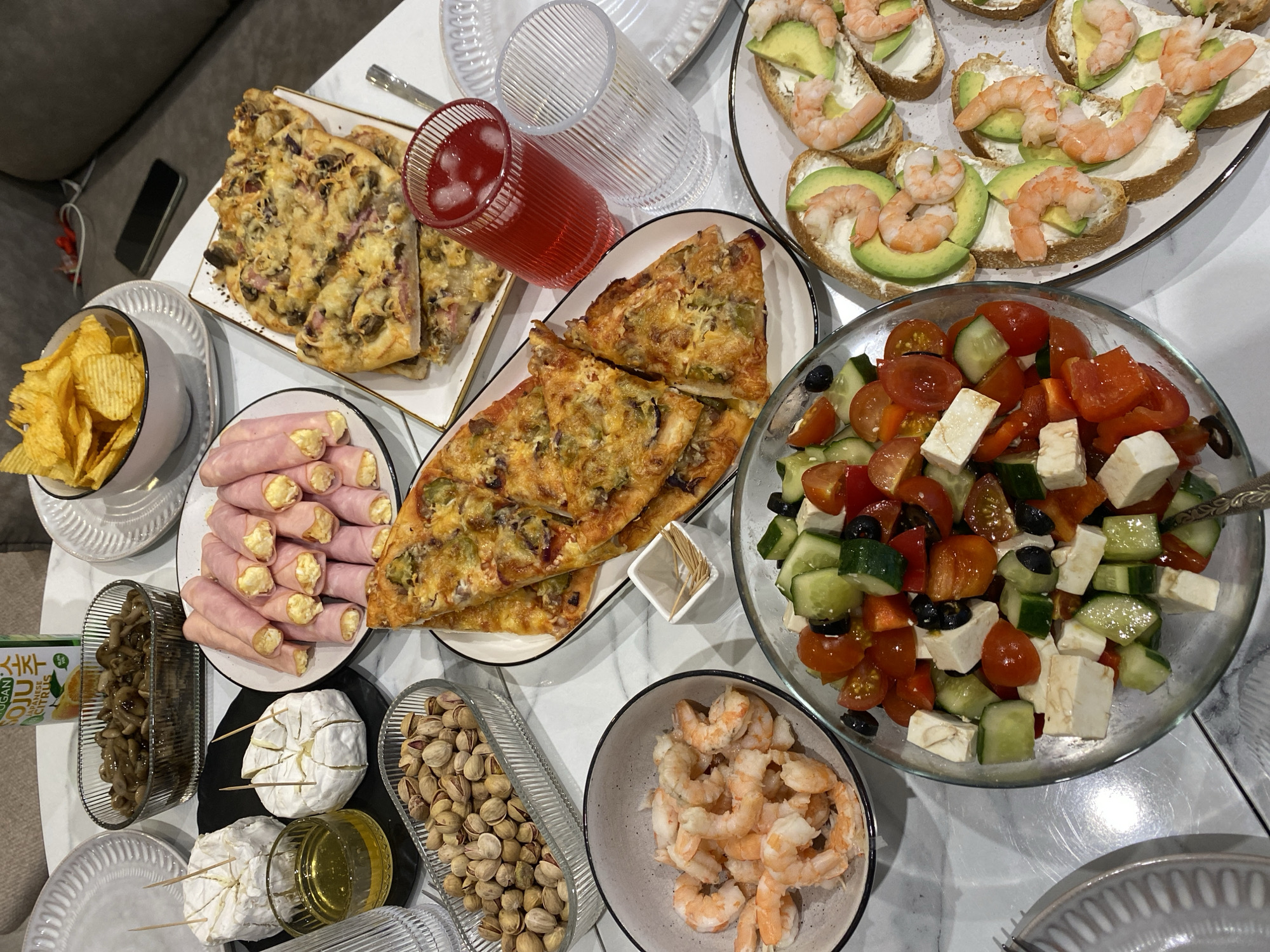 The image shows a tabletop spread of food items on a white marble surface. There is no indication of the location being Izhevsk, Russia or any people.
The food includes:
* Two plates of pizza, one with a rectangular cut and the other with triangular slices.
* A plate of ham rolls with a creamy filling.
* A bowl of potato chips.
* A plate of bread topped with cream cheese, avocado, and shrimp.
* A bowl of Greek salad with cucumbers, tomatoes, red peppers, olives, and feta cheese.
* A bowl of cooked shrimp.
* A container of pistachios.
* A container of marinated mushrooms.
* Two wheels of brie cheese with toothpicks.
* A small bowl of honey.
* Two glasses, one filled with a red liquid.
* Several empty plates.