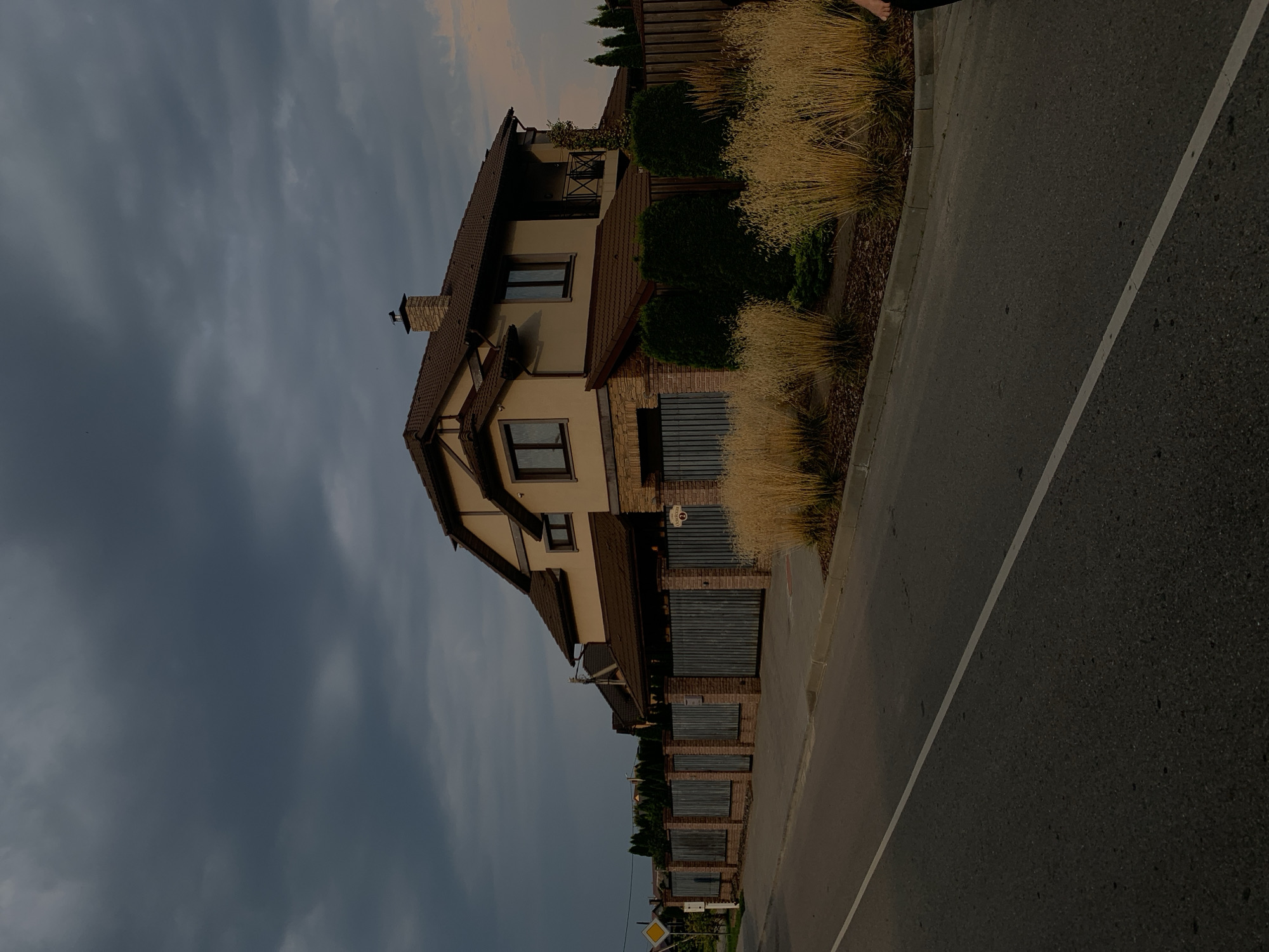 A two-story residential house with a brown tiled roof and beige exterior walls is situated behind a brick and metal fence. The fence has metal gates, one of which displays a house number. In front of the house, tall, golden-colored ornamental grasses are planted along a sidewalk that runs adjacent to a two-lane asphalt road. The sky is overcast with dark, heavy clouds. A yellow diamond-shaped sign with a black border and a black arrow pointing right is visible on the left side of the road. The scene appears to be in a suburban area. The image does not contain any discernible people or animals.