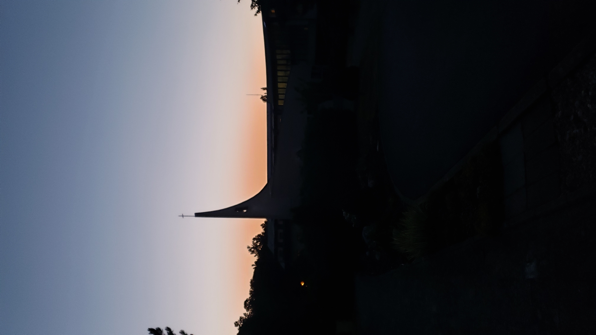 An external, ground-level, vertical shot captures a modern church with a striking curvilinear roofline and a prominent cross atop its spire, silhouetted against a dusky sky. The church, located in the city of Prešov, Slovakia, is surrounded by dark foliage. The foreground is a paved area, likely a road or pathway, leading towards the church. The time of day appears to be dusk or dawn, as indicated by the gradient of the sky, transitioning from a deep blue at the top to a pale orange hue near the horizon. There are no people visible in the image. The lighting conditions are low, with the church and surrounding landscape rendered in shadow.