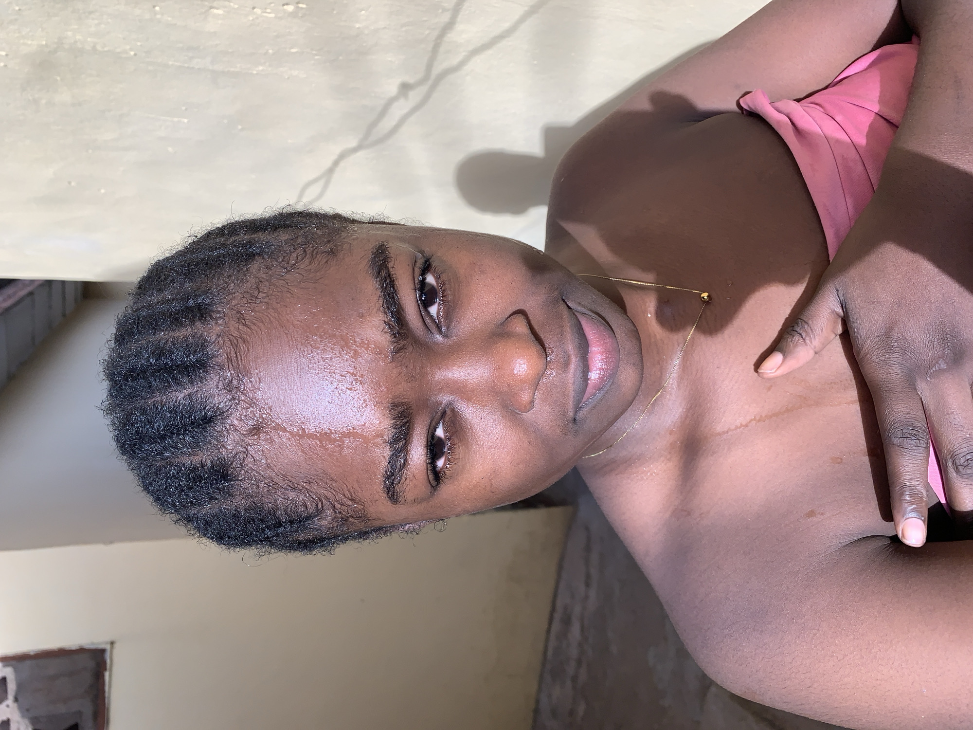 A medium-shot image shows a dark-skinned individual, possibly female, with short, braided hair appearing wet. The person is wearing a pink top and has a gold necklace. The background consists of a plain, light-colored wall with a visible crack and some shadows. The location is undetermined. There are no indicators to confirm location within Rafin Par, Nigeria. The lighting appears to be artificial. The individual is looking directly at the camera. Their hands are partially visible.