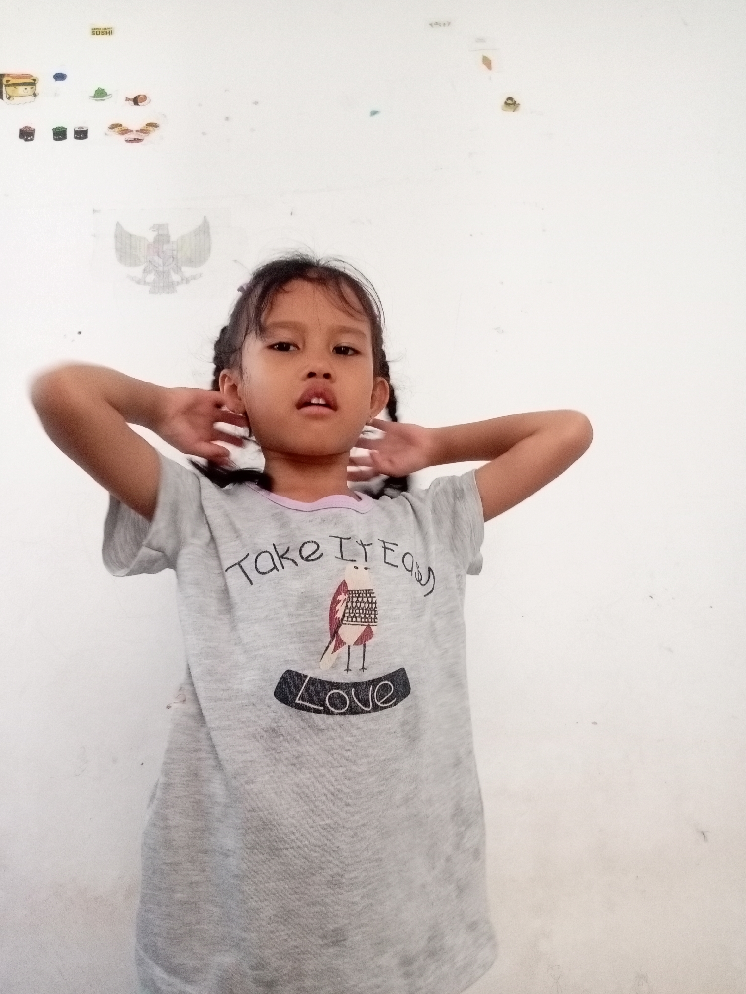 A young girl with dark hair in braids stands against a white wall. She is wearing a light gray t-shirt with a graphic design of a bird and the word "Love" printed on it. Her hands are behind her head, and she is looking towards the camera. The wall behind her has several colorful stickers, including images of sushi and an eagle. The overall scene suggests an indoor setting, likely a home or classroom, given the decorations on the wall. The girl's pose and direct gaze indicate she is aware of being photographed.