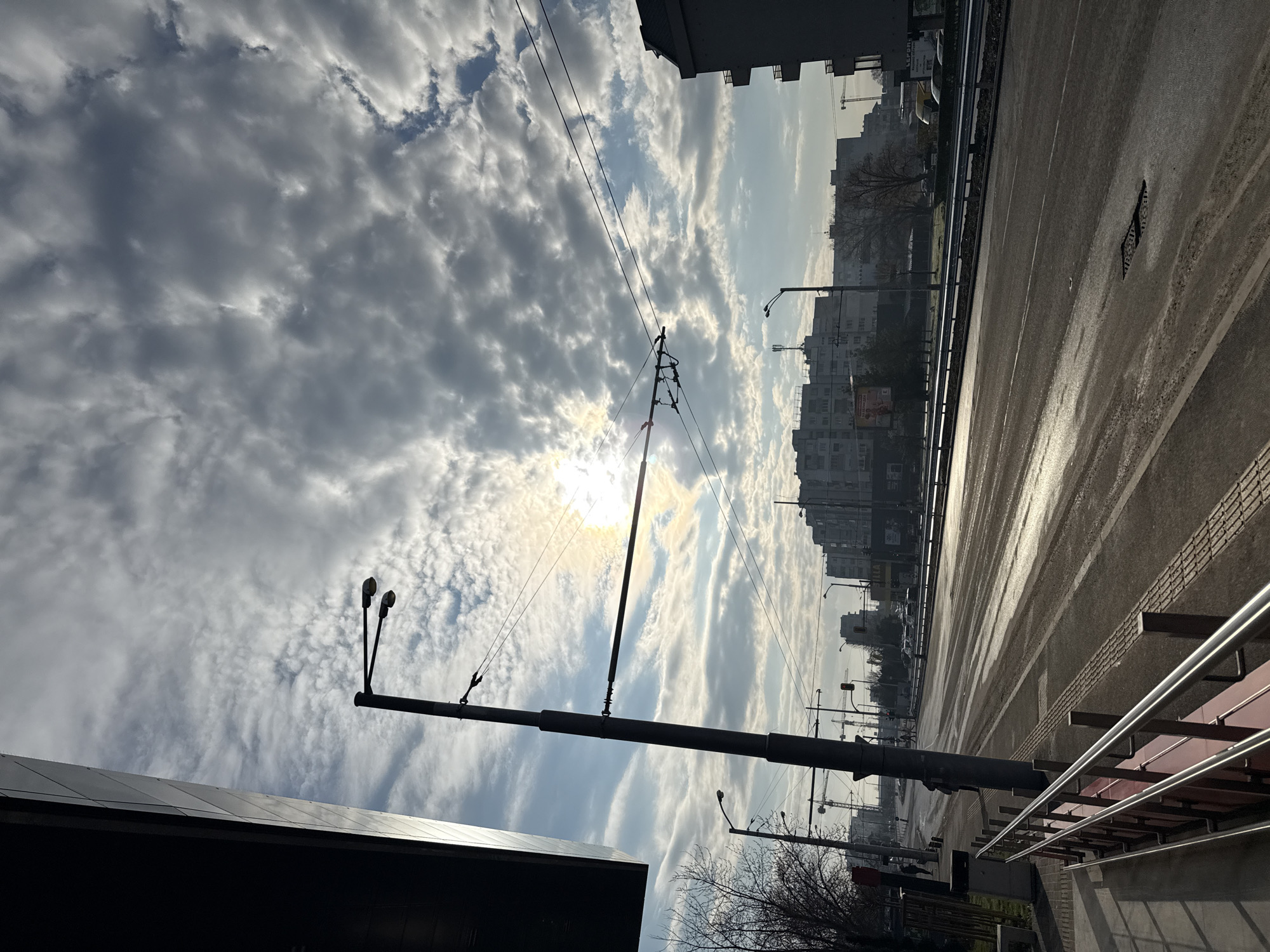 An eye-level, rotated image depicting a sun-lit urban street scene in Sofia, Bulgaria. The sky is filled with billowing white and gray clouds partially obscuring the sun. A streetcar pole and wires are prominently placed in the foreground, silhouetted against the bright sky. A dual-lamp street light extends from the pole. The background contains apartment buildings, more street lights, and utility poles lining both sides of a wide, multi-lane road. A metal railing runs along the right side of the frame next to a set of stairs. The street appears wet with some puddles reflecting light. The overall color scheme is dominated by shades of gray, white, and blue.