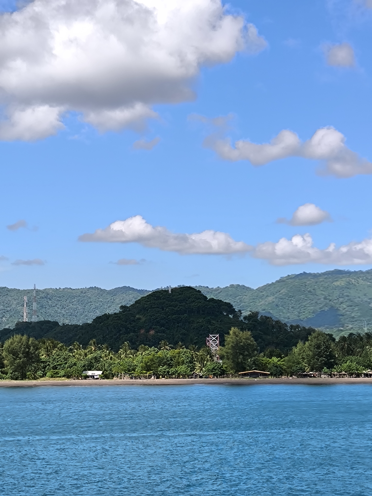 This vertical shot captures a vibrant coastal landscape in Lembar Selatan, Indonesia, under a bright, sunny sky. The foreground is dominated by calm, clear blue-green water, showing subtle ripples across its surface.
A dark, narrow sandy beach forms the shoreline, separating the water from the land. Along the beach, several small, simple wooden huts or buildings with dark roofs are nestled amongst dense tropical vegetation. This vegetation, comprising various trees including palm trees and what appear to be pines, creates a lush green border along the coast.
Behind the immediate beachfront, a prominent, verdant hill rises, covered in dense, dark green foliage. On top of this central hill, a distinct white structure, possibly a cross or monument, stands out. To the left, a tall, slender communication or power tower is visible on a distant, forested hill, while another, larger tower with an 'X'-patterned structure can be seen further to the right, partially obscured by trees near the beach. In the background, layers of more forested hills and mountains extend under the bright blue sky, which is adorned with scattered, fluffy white cumulus clouds.
The scene depicts a serene, natural environment during daytime, likely mid-morning or afternoon, characterized by clear weather and abundant sunshine. No human activity or visible text is present in the image.