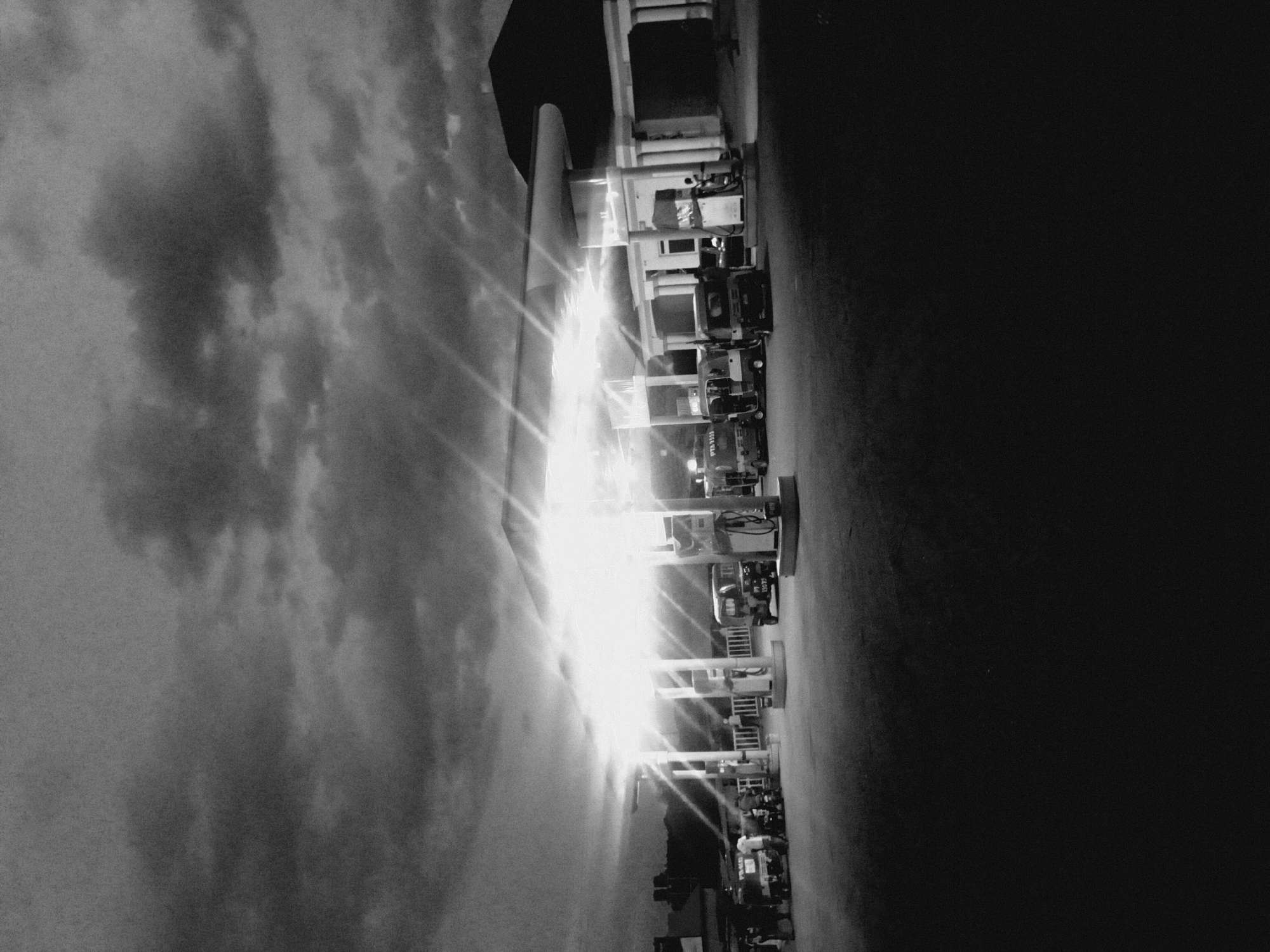 The image shows a nighttime scene in Bukuru, Nigeria. A brightly lit gas station is prominent, with multiple pumps and what appear to be parked vehicles. A building is adjacent. The sky above the gas station is dark, with cloud cover visible. To the far right, the image fades to black with few distinguishing features. There are blurred people at the gas station.
