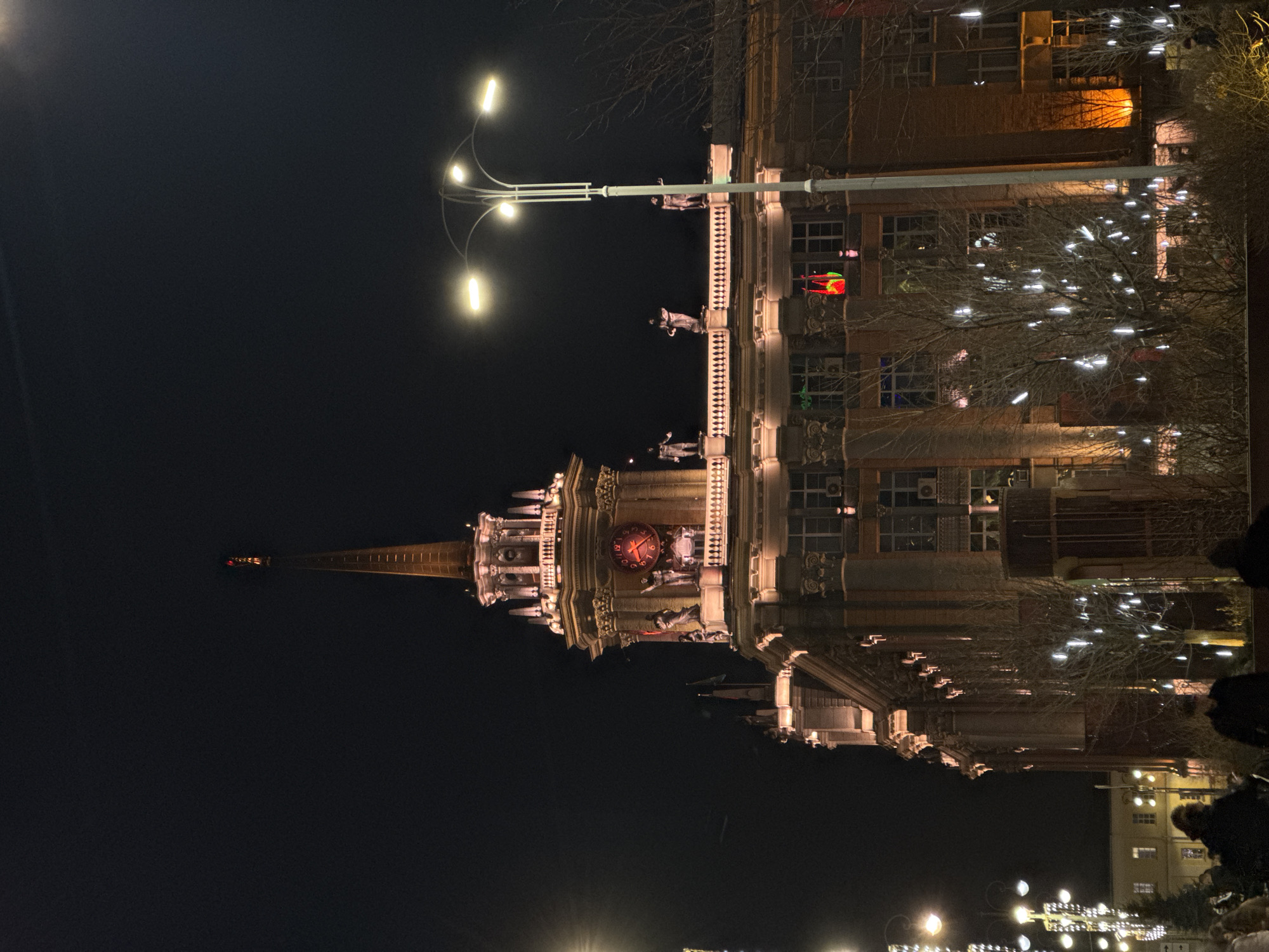 Here is a forensic description of the image.
The image depicts a tall building with a prominent clock tower at night. The building is illuminated with warm lighting, highlighting its architectural details. A streetlight with multiple bulbs is visible, casting light on the building's facade. The tower's clock face glows red. Bare branches with decorative white lights are visible to the right of the building. At the bottom right corner of the image, blurred figures of people can be seen. The sky is dark, indicating a nighttime setting. The location appears to be an urban area, possibly a city center.