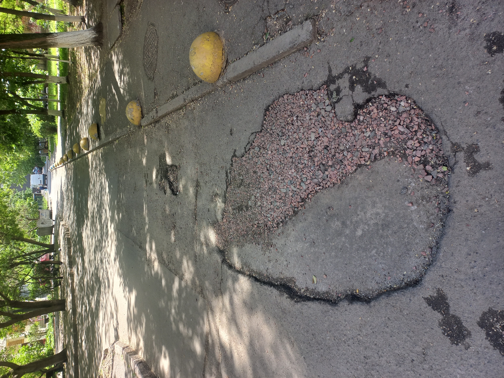 This image depicts a damaged asphalt road stretching into the distance, with a prominent, large pothole on the right side of the frame. The pothole is irregularly shaped and filled with reddish-brown gravel, highlighting the significant deterioration of the road surface. Its edges are rough and uneven.
To the left of the road, a row of yellow, dome-shaped concrete bollards runs parallel to a concrete curb. These bollards show signs of wear and chipped paint, forming a barrier between the road and a grassy strip populated by mature trees with green foliage. Strong sunlight casts distinct, dappled shadows from the trees and bollards across the asphalt, indicating a bright daytime setting, likely late morning or early afternoon. A metal manhole cover is visible near the curb further up the road. In the distance, between the trees on the left, a white van is partially visible, suggesting an urban or suburban setting. The scene conveys a public outdoor space, possibly a street or a path, experiencing disrepair. No discernible text is visible in the image.