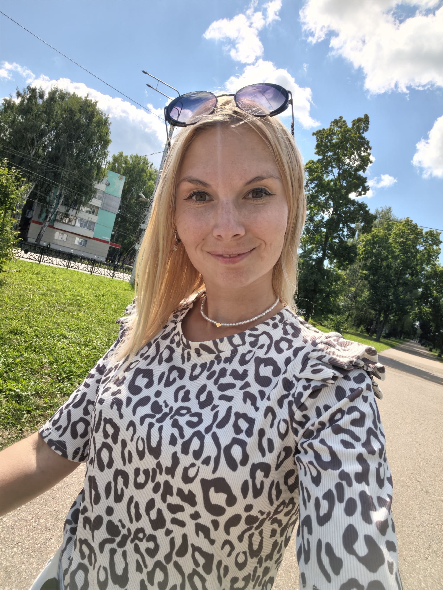 A blonde woman with sunglasses perched on her head smiles at the camera. She is wearing a leopard-print top and a pearl necklace with a small gold heart pendant. In the background, a green lawn, trees, a building, and a paved road are visible under a blue sky with white clouds. The scene appears to be outdoors in an urban or suburban setting, possibly in Sterlitamak, Russia, given the context. The lighting suggests it is daytime.