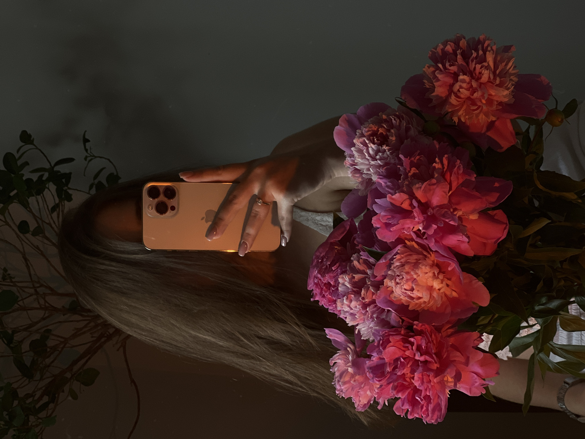 The image depicts a person holding a smartphone in their right hand, partially obscuring their face. The smartphone, an iPhone, is gold-toned and features a prominent camera array. The person's hand has manicured nails, and a ring is visible on their ring finger. They are wearing a white garment with textured detail. In the foreground, a large bouquet of vibrant pink peonies is arranged, with some flowers appearing fully bloomed and others in bud. The background consists of a dark, textured wall and a shadowy plant with delicate branches and leaves. The lighting suggests an indoor setting, possibly a home environment in Ternopil, Ukraine, with warm light illuminating the peonies and casting shadows. The overall mood is intimate and artistic.