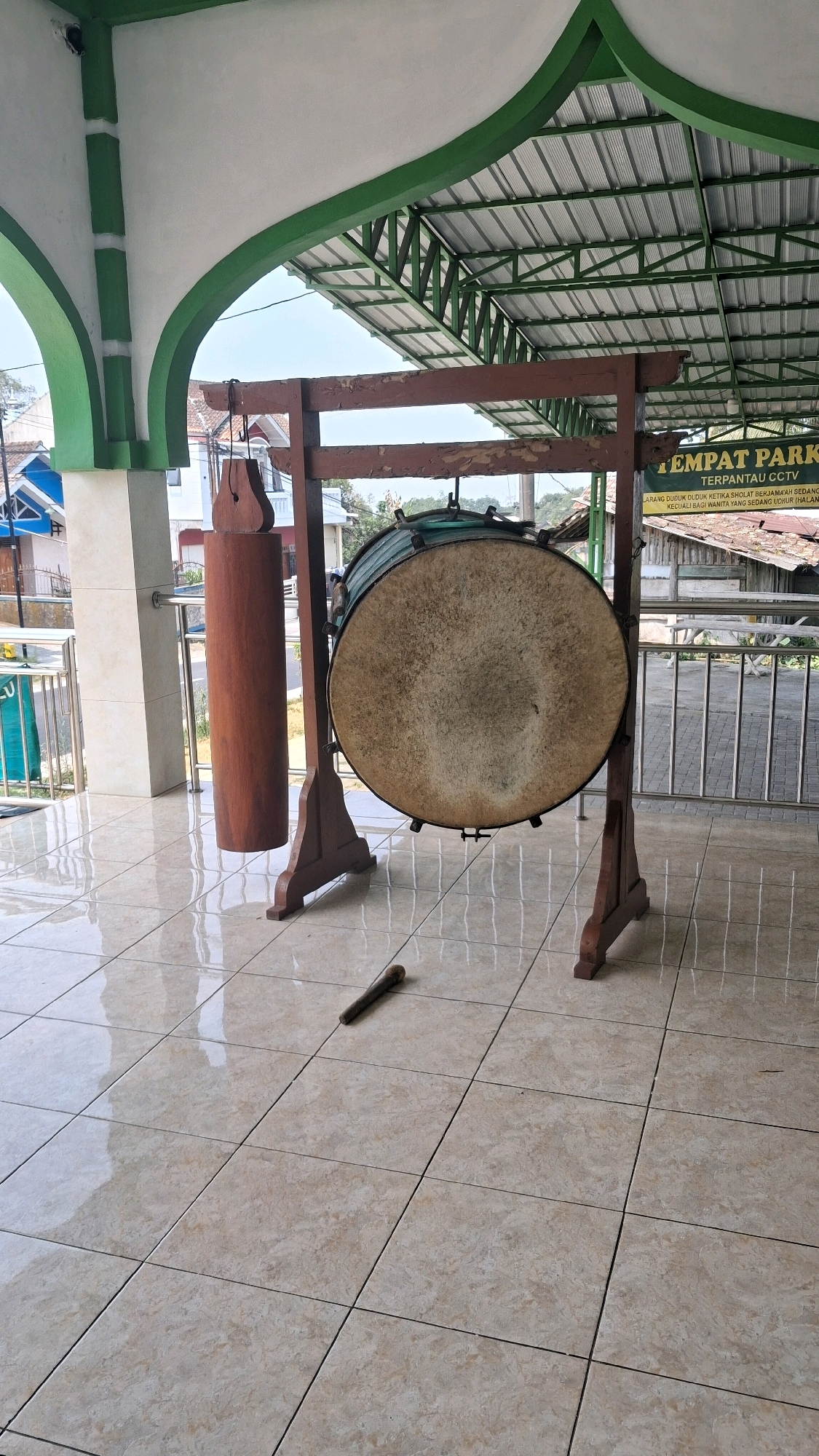 The image depicts a large, round drum with a textured surface, likely made of animal hide, suspended on a wooden frame. To the left of the drum, a smaller wooden object, possibly a mallet or a decorative piece, hangs from a rope. On the tiled floor in front of the drum, a wooden stick lies horizontally. The drum and its frame are situated outdoors, under a green and white arched roof structure, supported by columns. In the background, a metal fence with railing is visible, separating the area from a street and buildings. A sign with Indonesian text, including "TEMPAT PARKIR" (parking lot) and "TERPANTAU CCTV" (under CCTV surveillance), is also present in the background. The overall setting suggests a public space, possibly associated with a mosque or community building in Giriwarno, Indonesia.