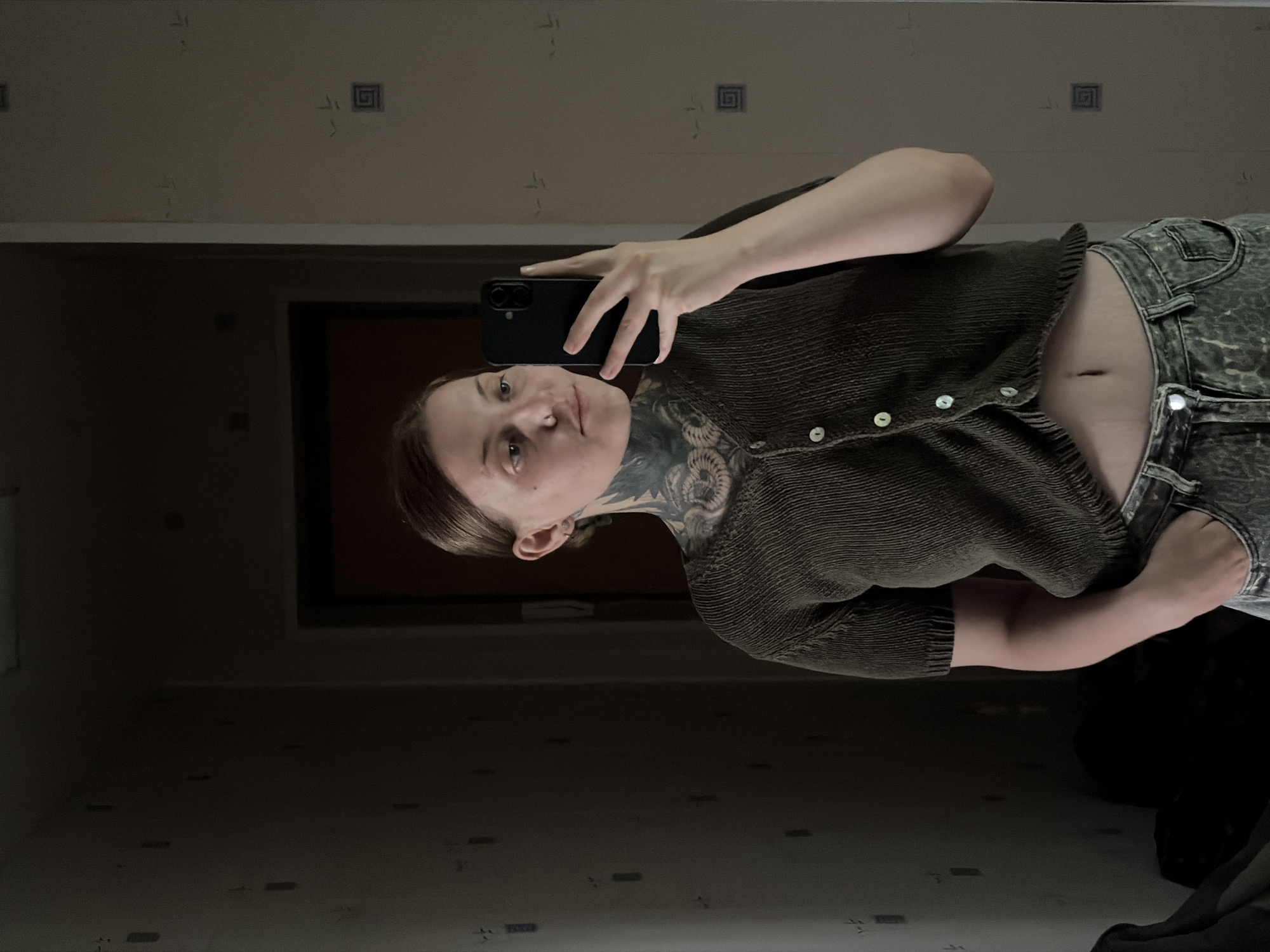 A person is taking a selfie in a mirror. The person is wearing a dark green, ribbed knit top with a button-down front and acid-wash jeans. The top is cropped, exposing a portion of their stomach. The person has dark hair pulled back and is looking at the camera. They have extensive dark tattoos visible on their neck and chest.
The setting appears to be a room with light-colored walls, possibly a bathroom or bedroom, with a large mirror. The mirror reflects a dark doorway or closet in the background. The lighting is dim, suggesting it might be nighttime or a poorly lit room. There are some small, dark rectangular objects scattered on the floor in the reflection.
The image is taken vertically. The person is holding a black smartphone in their right hand, angled to capture the selfie. The overall mood is casual and personal. No text is visible in the image.