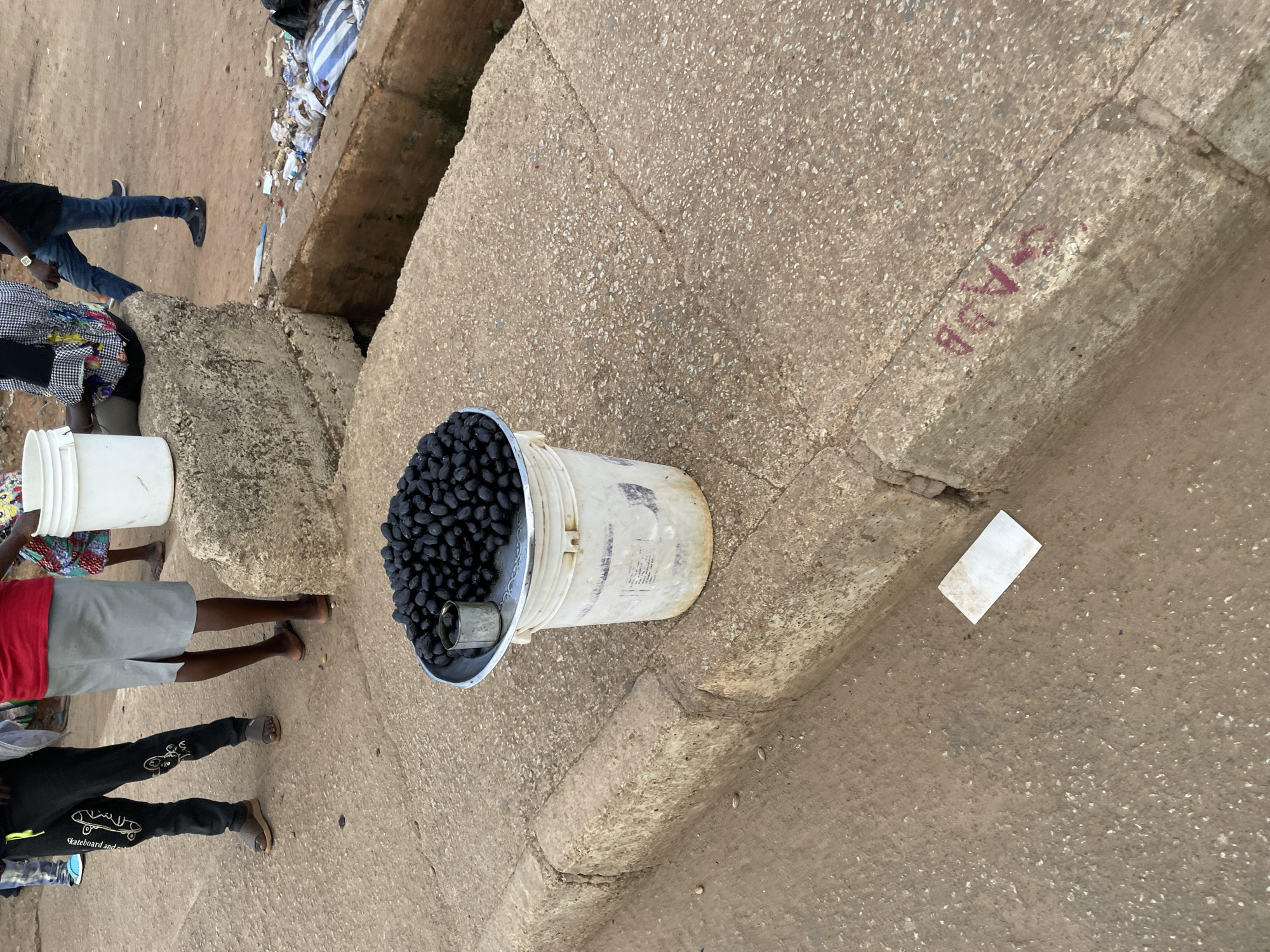 An outdoor, eye-level, medium shot shows a bucket of black olives on a curb in what appears to be a city. A person on the left is wearing black pants with white graphics and sandals. Another person in the center is wearing a red shirt and a grey skirt. A third person on the right is wearing blue jeans and black clogs. A white bucket is placed on the curb with a silver tray on top filled with black olives. A small metal can is in the tray. The ground is made of concrete and dirt. There is a drainage ditch to the right of the curb. Trash is visible in the ditch. A white piece of paper is on the ground in front of the curb. Red graffiti is on the side of the curb.