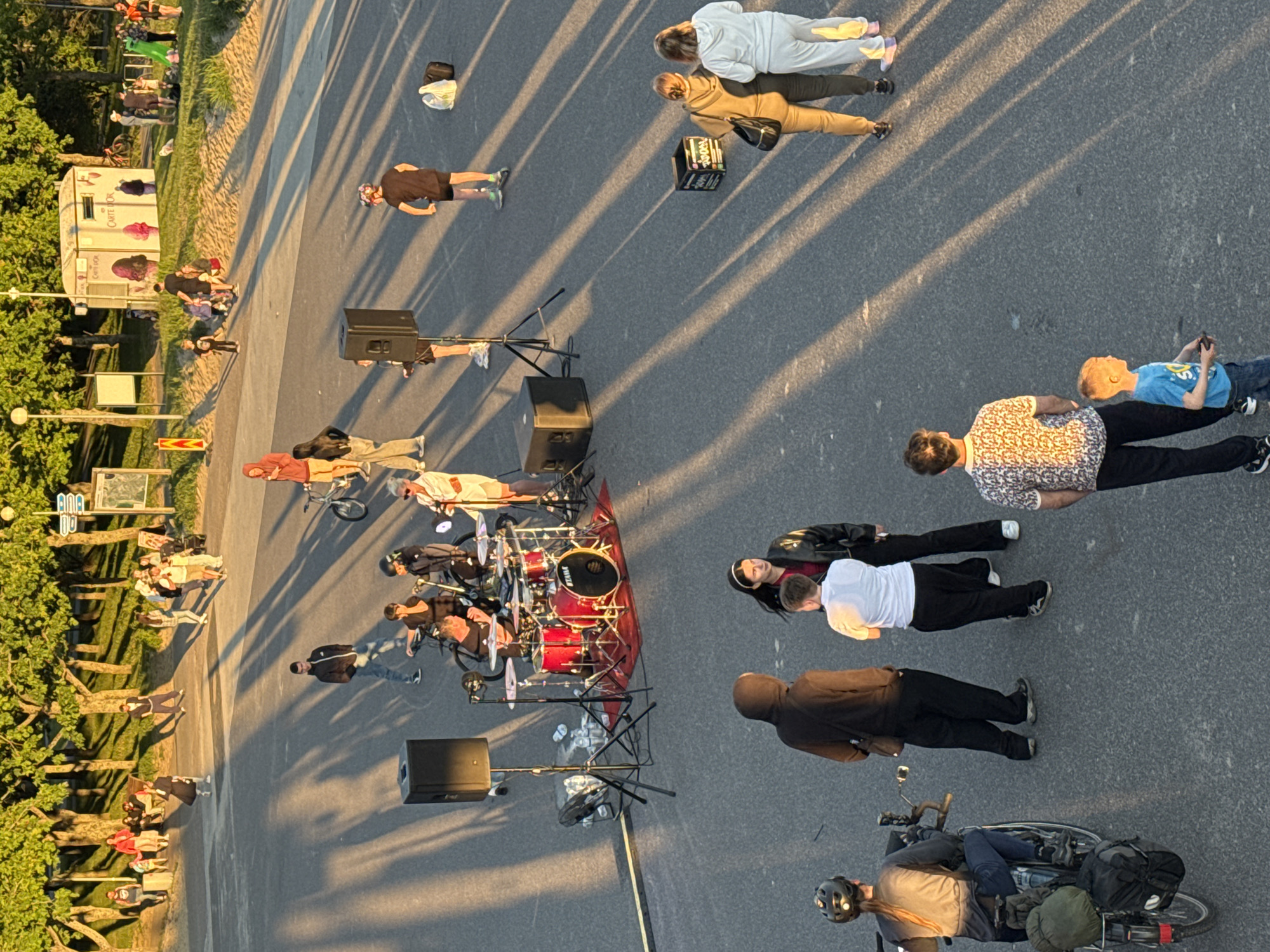 The image captures an outdoor scene in Tallinn, Estonia, likely during a sunny afternoon or early evening, as indicated by the long shadows cast across the asphalt. In the foreground, a person on a bicycle with loaded panniers is positioned to the left, facing away from the viewer. A group of individuals is gathered near a drum set set up on a red rug, with a large speaker and other musical equipment nearby. Several people are standing or sitting around the musical setup, some appearing to be musicians or audience members. Further back, a diverse crowd of people are seen walking, sitting, and interacting in a park-like area with trees lining the background. A small, light-colored booth with signage is visible in the mid-ground, possibly a food or information stand. The overall atmosphere suggests a public gathering or event taking place.