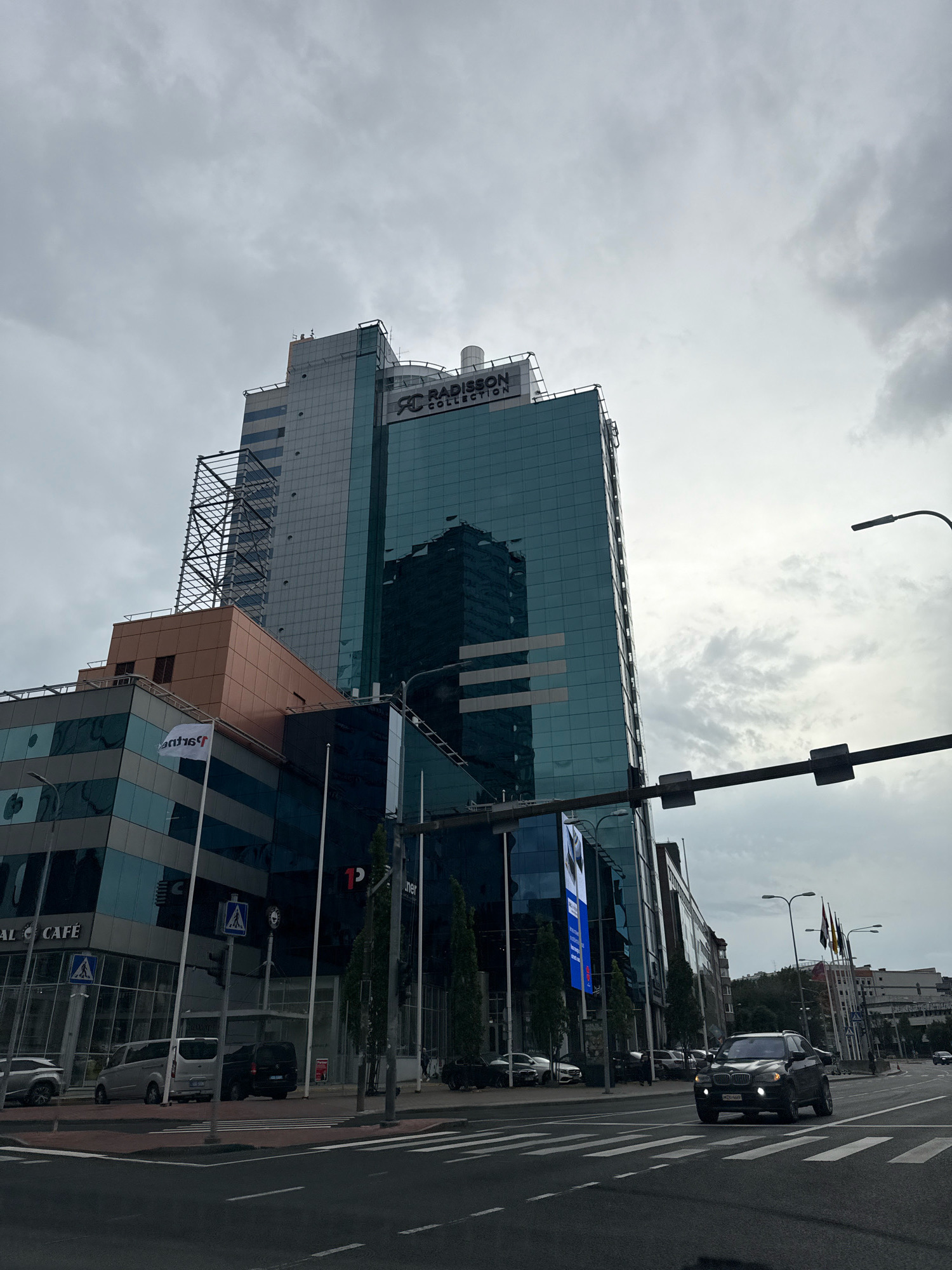 An overcast day in Tallinn, Estonia, captures a view of the Radisson Collection Hotel, Tallinn, a modern skyscraper with a reflective glass facade. The building's prominent signage, "RADISSON COLLECTION," is visible near its apex. In the foreground, a street intersects with a pedestrian crossing. Several vehicles are present: a dark SUV driving towards the viewer, a silver van parked on the side of the road, and other cars further in the distance. A few trees line the sidewalk. Flags of various nations are displayed on poles in the background, adjacent to other buildings. The overall scene is one of urban infrastructure under a grey sky.
