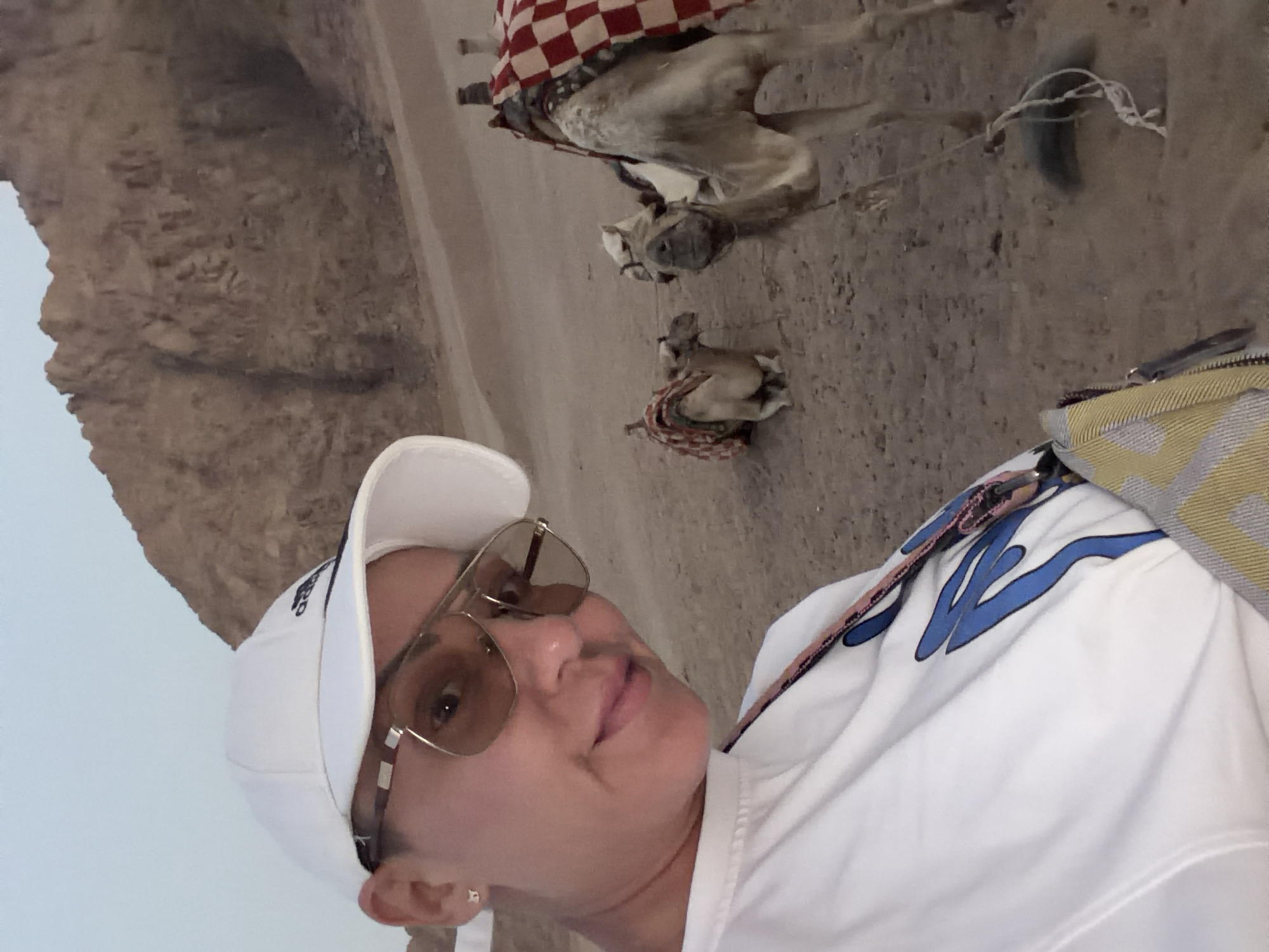 A woman wearing a white baseball cap and sunglasses takes a selfie in a desert landscape. She is wearing a white t-shirt with blue lettering. In the background, two camels are resting on the sandy ground. One camel is closer to the camera and has a red and white checkered blanket on its back. The other camel is further away and also has a blanket. The terrain is sandy with rocky hills in the background, suggesting a desert environment. The sky is a pale blue. The location is identified as Sharm El Sheikh, Egypt.