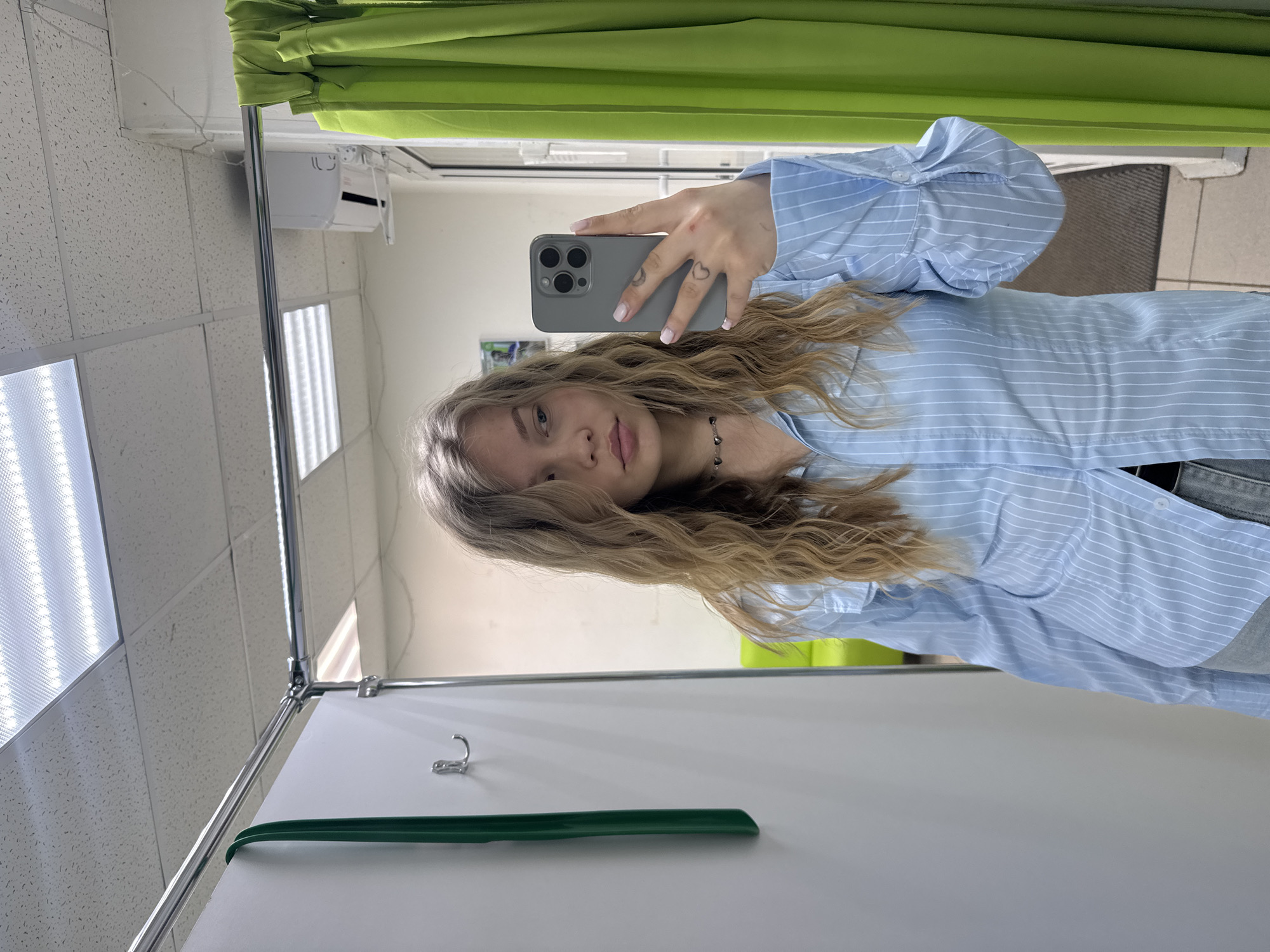A person stands in what appears to be a fitting room, taking a selfie with a gray smartphone. The individual, likely female, has blonde wavy hair and is wearing a light blue striped shirt. The phone in hand partially obscures the face. Visible on the hand holding the phone are small heart tattoos.
The fitting room has a green curtain at the top of the frame. Fluorescent lighting fixtures are visible in the ceiling. A white partition wall is in the foreground, along with a green clothes hanger resting against it. A metal rack with a hook is positioned near the top of the wall.
Based on geolocation information, the location is Chaikovsky, Russia.