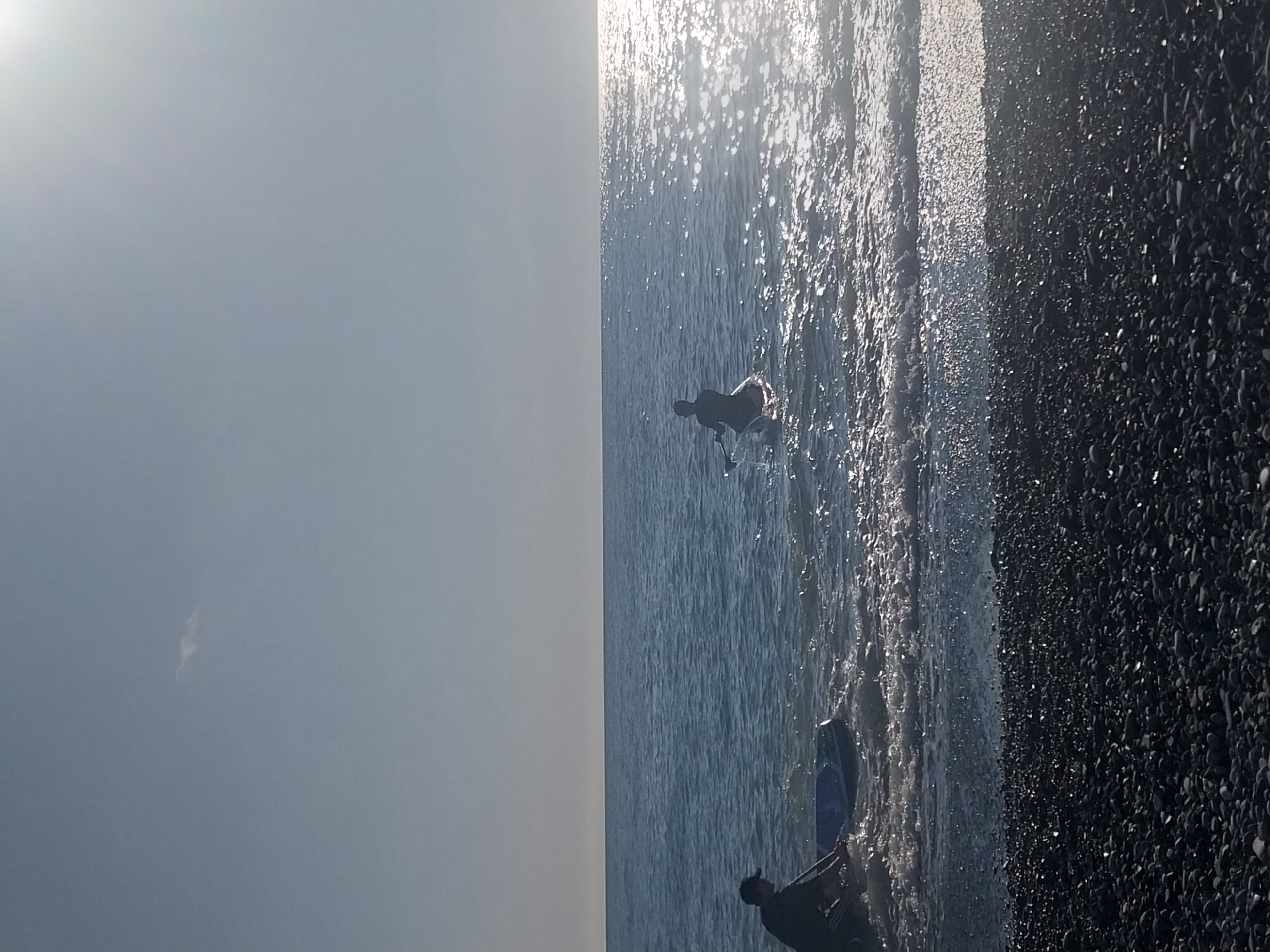 The image captures a sunlit beach scene in Sochi, Russia, with two individuals in the water. The photograph is oriented vertically, with a bright, largely clear sky occupying the left side. The middle section is dominated by the sea, glistening intensely with sun glare, creating a sparkling, reflective surface. To the right is a narrow strip of dark pebble beach, densely covered with small, smooth stones.
Two people are visible as dark silhouettes in the water. One person is positioned closer to the foreground within the water section, partially submerged and seen from behind, appearing to be sitting or kneeling. Further out in the water, another individual is also partially submerged and appears to be bending forward or interacting with the water.
The time of day appears to be mid-day or early afternoon due to the strong, overhead sunlight causing significant glare on the water. The weather is clear and bright, indicated by the uniform light grey sky. The scene depicts a leisure activity, likely people enjoying the sea on a sunny day at the Black Sea coast. There is no visible text in the image.