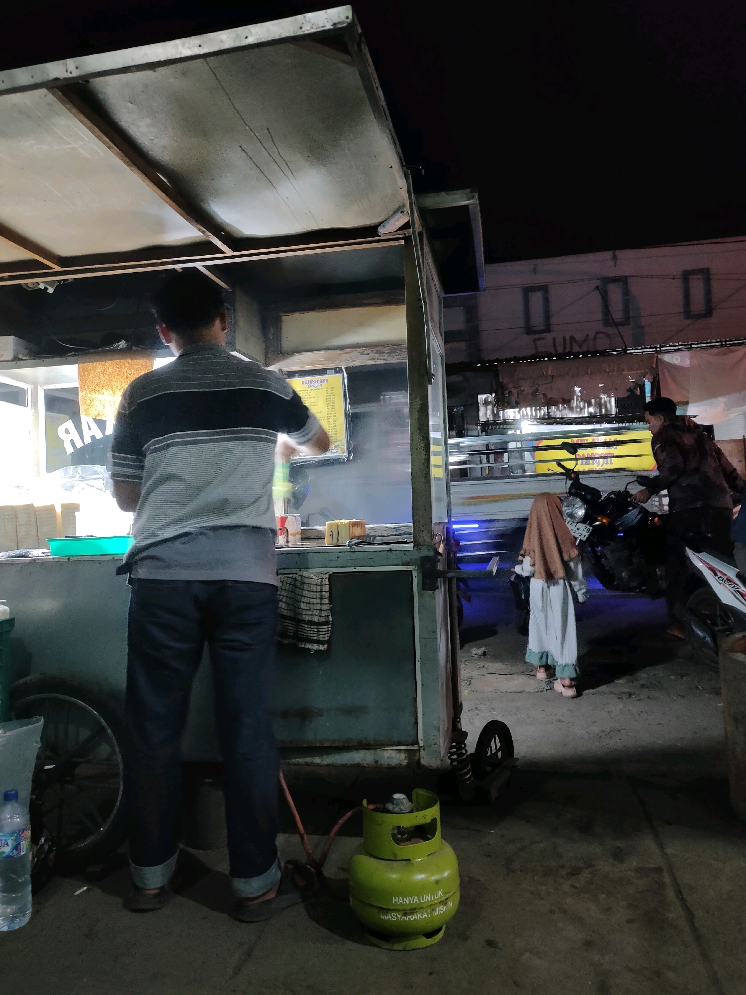 The image depicts an outdoor scene at night, likely in Codo, Indonesia, focused on a street food stall. A man, facing away from the camera, is preparing food behind the stall. He is wearing a grey and black striped short-sleeved shirt and dark pants. In front of the stall, a green LPG gas cylinder with text indicating it is for the poor is visible. To the right of the stall, a motorcycle is parked, with a person in a brown jacket and a young child wearing a light brown hijab and a light-colored dress standing near it. The background shows a building with dimly lit windows and some signage. The ground is paved, and there is a water bottle near a cart wheel on the left side of the image. The overall lighting suggests it is nighttime, with artificial light sources illuminating the immediate area of the food stall.