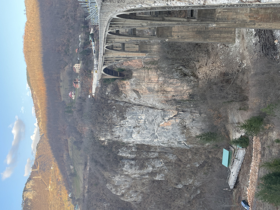 The image displays the Đurđevića Tara Bridge, a multi-arch concrete structure, spanning a deep river gorge in Đurđevića Tara, Montenegro. The bridge extends from the right foreground into the upper center, showcasing multiple supporting arches. The gorge walls consist of steep, rocky cliffs with varied grey and reddish-brown hues, sparsely covered by leafless deciduous trees and scattered evergreen conifers. At the base of the gorge on the left, a wooden platform supports a small building with a green roof, beside which a dark-colored sedan is parked on a dirt patch. Beyond the gorge, forested mountains rise, with the upper slopes of the mountain on the left receiving direct sunlight, casting an orange hue, while other slopes remain in shadow. Several small residential buildings with red roofs are visible on the distant hillsides. The sky above is clear blue with a few scattered white clouds. No individuals or specific actions are discernible within the frame.