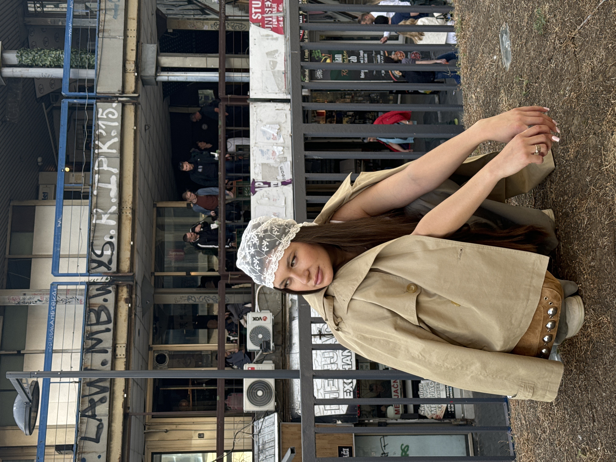 The image shows a woman in a tan trench coat and lace bonnet kneeling in front of a wrought iron fence. Behind the fence is a street scene in Belgrade, Serbia. There is graffiti on the buildings and people are visible in the background. There are air conditioning units attached to the building in the background. The signage visible includes advertisements for stores and restaurants. The woman is looking at the camera and her hands are clasped in front of her.