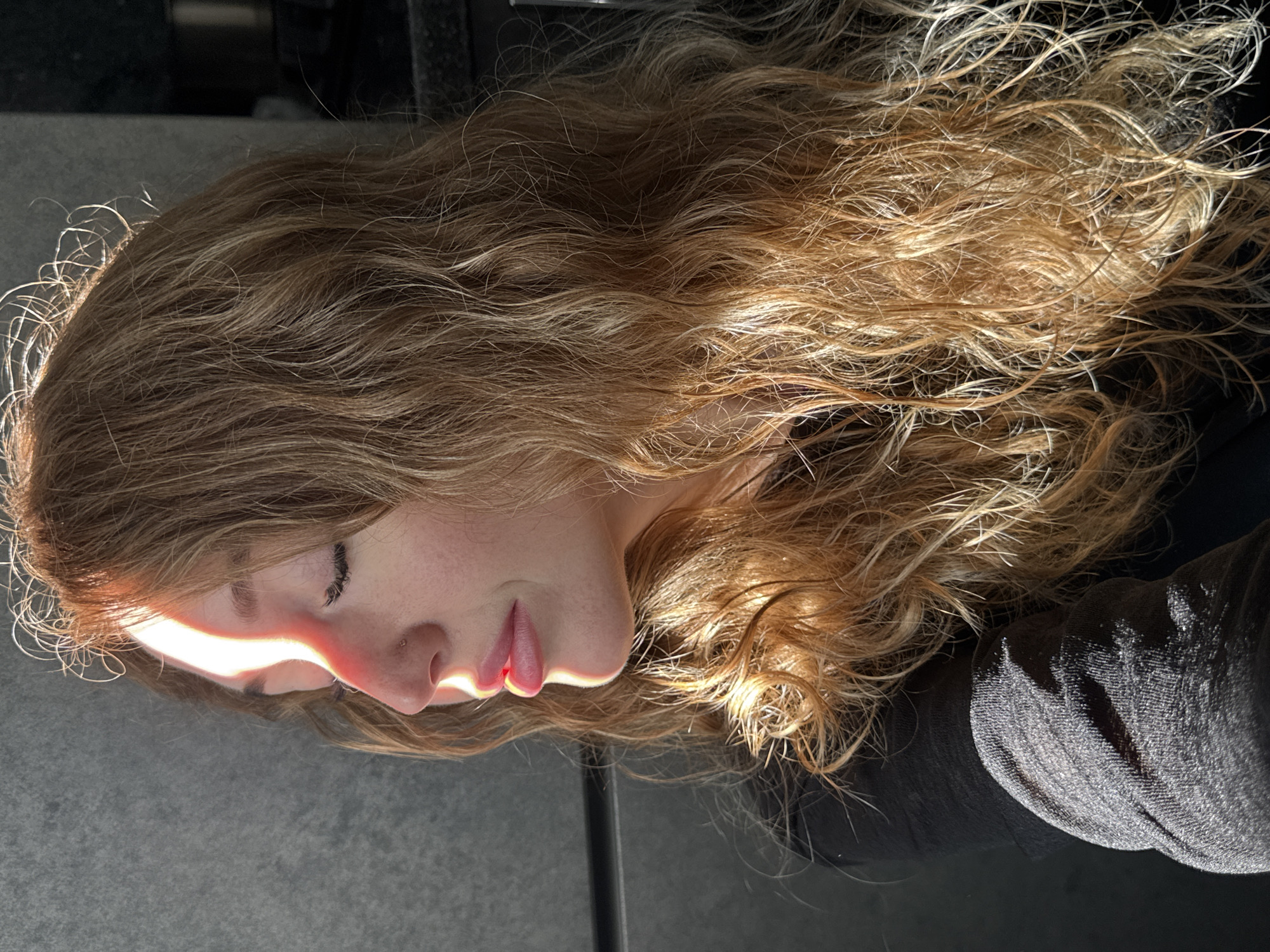 An outdoor, medium shot shows a young woman with light brown, wavy hair in profile, facing left. She is wearing a black top. The woman's eyes are closed, and a bright beam of sunlight illuminates the left side of her face, from her forehead down to her lips, creating a sharp contrast with the shadowed right side of her face. The background is dark and appears to be a textured, grey wall on the left and a darker surface on the right, possibly a kitchen counter and cabinets. The image does not contain enough information to determine the specific location in Chaikovsky, Russia.