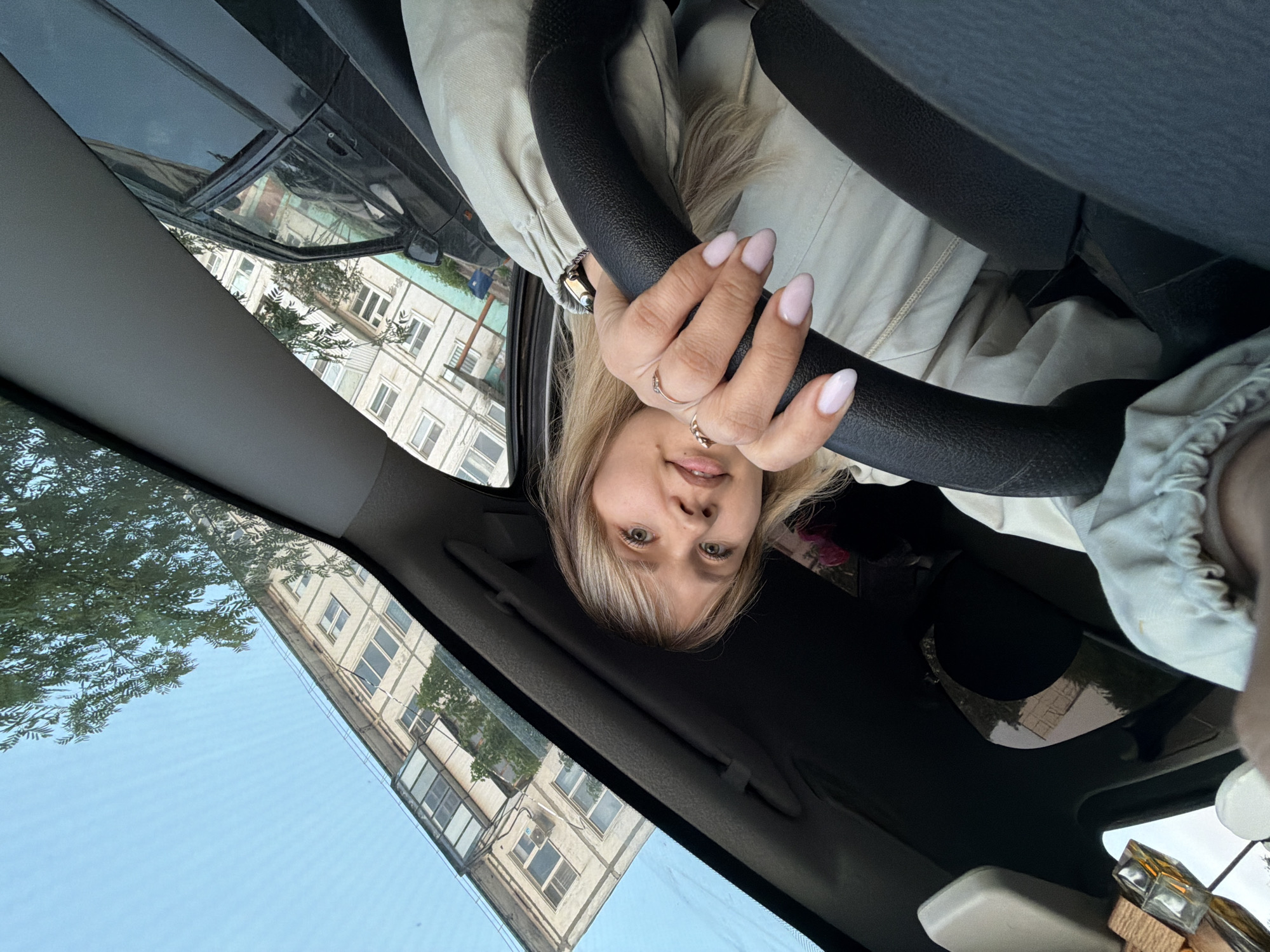 A woman is sitting in the driver's seat of a car, holding the steering wheel. The car is parked near an apartment building. The apartment building is likely located in the city of Chaikovsky, Russia, given the background of the image. The woman is wearing a light-colored jacket and has blond hair. Her nails are manicured. The sky is visible through the car's windows.