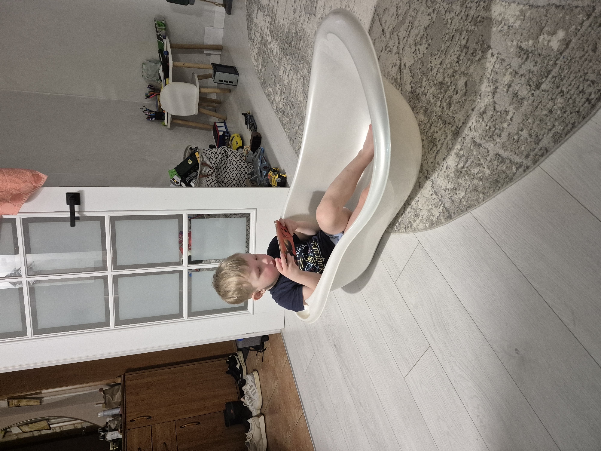 A young male child with blonde hair, wearing a dark t-shirt and light shorts, sits barefoot inside a white, freestanding bathtub positioned on a light wood-grain laminate floor. He holds a red-backed smartphone in both hands, gazing down at its screen. Behind the child is a white door with multiple glass panes, open to reveal two adjacent rooms. To the left, through the doorway, a room with darker wooden flooring shows a brown wooden cabinet topped with items and a mirror above it. Several pairs of sneakers are visible on the floor near the cabinet. To the right, another room with light grey walls contains a small white desk, a white chair, various writing utensils, a woven basket filled with toys, and several toy vehicles scattered on a grey patterned rug. An orange towel is draped over the upper right corner of the door frame. The scene is set indoors within a residential environment in Chaikovsky, Russia.
